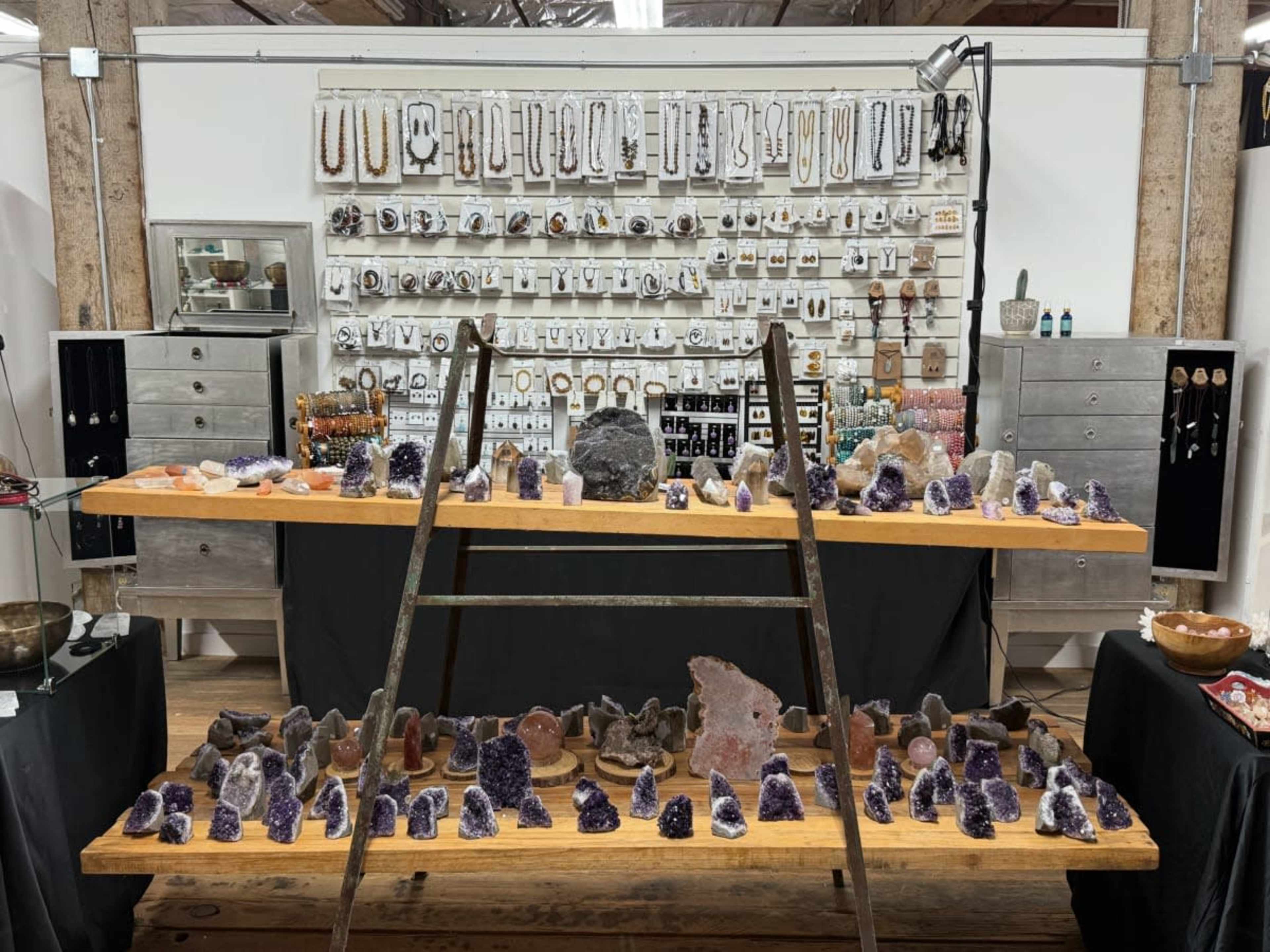 A display of various gemstones and jewelry is arranged on multiple wooden tables, with additional items hanging on a wall behind them.