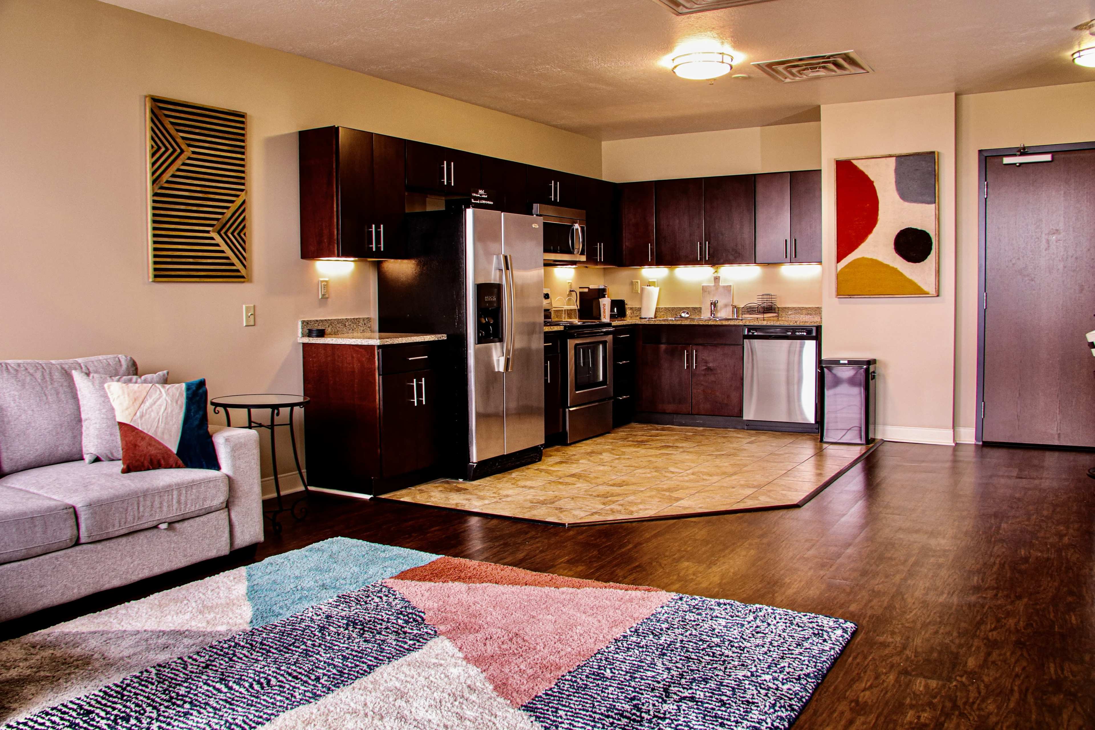 The image shows a modern kitchen with stainless steel appliances, dark wooden cabinets, and a cozy living area featuring a light gray sofa and a colorful area rug.