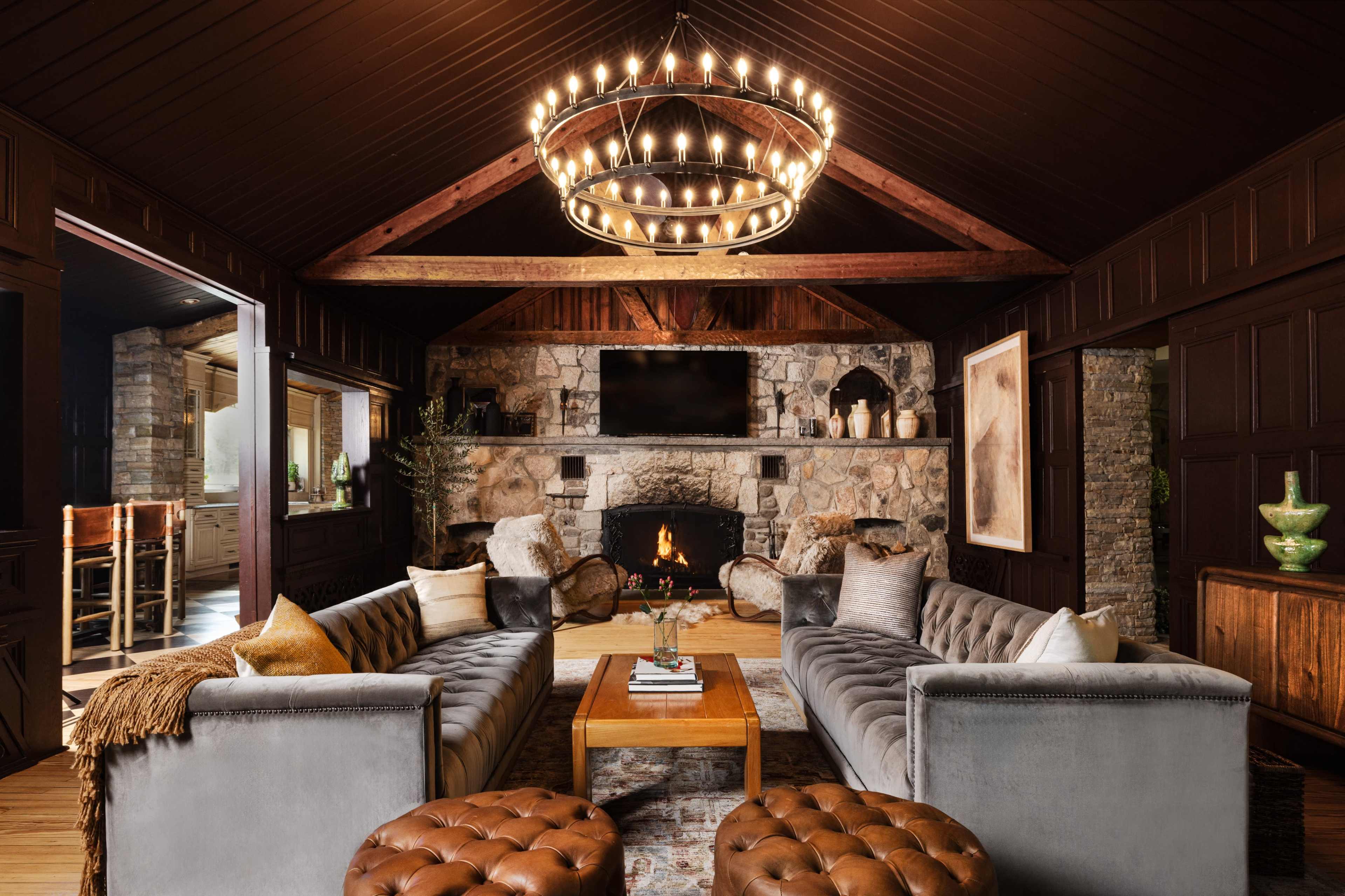 A cozy living room features two velvet sofas facing a stone fireplace, with a large chandelier overhead and wooden beams framing the ceiling.