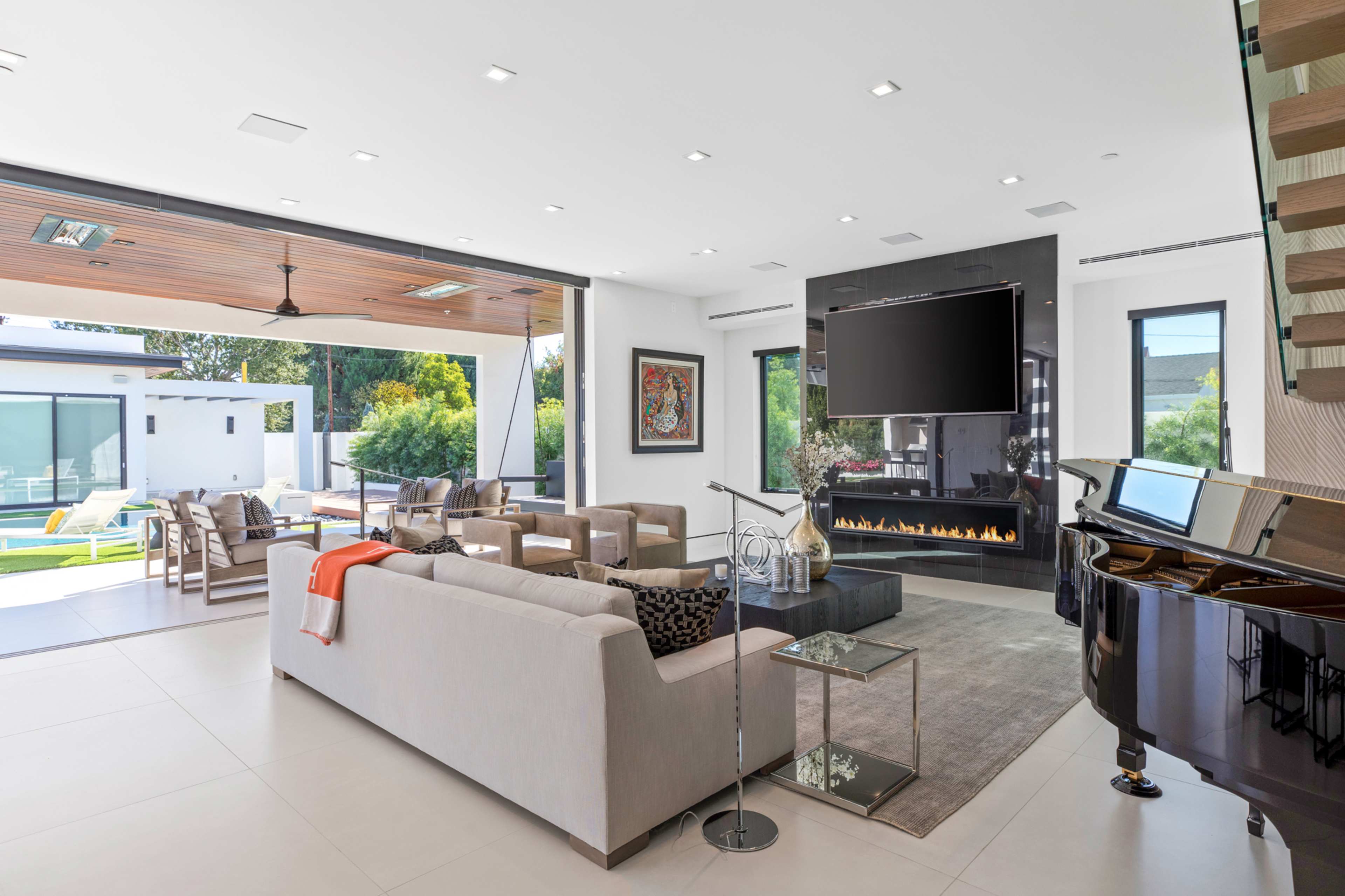 A modern living room features a light-colored sofa, armchairs, a sleek fireplace, and a grand piano, with large windows opening to an outdoor patio.