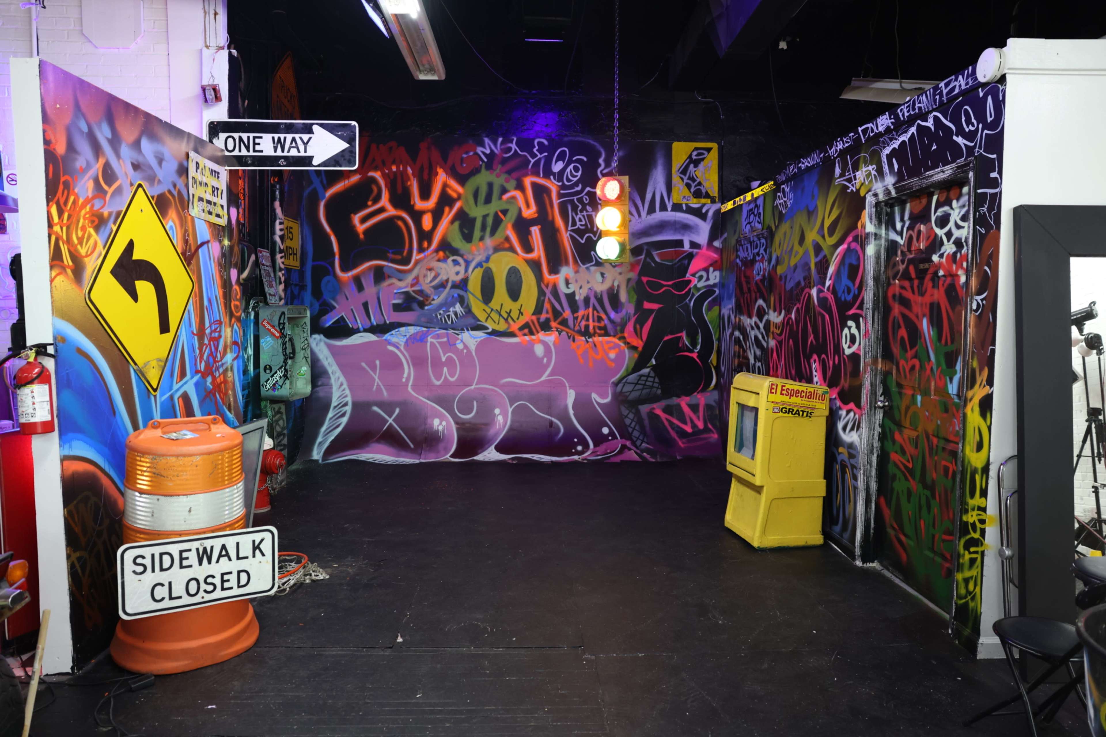 The image shows a vibrant urban scene with colorful graffiti-covered walls, street signs, and a yellow utility box in a dimly lit space.