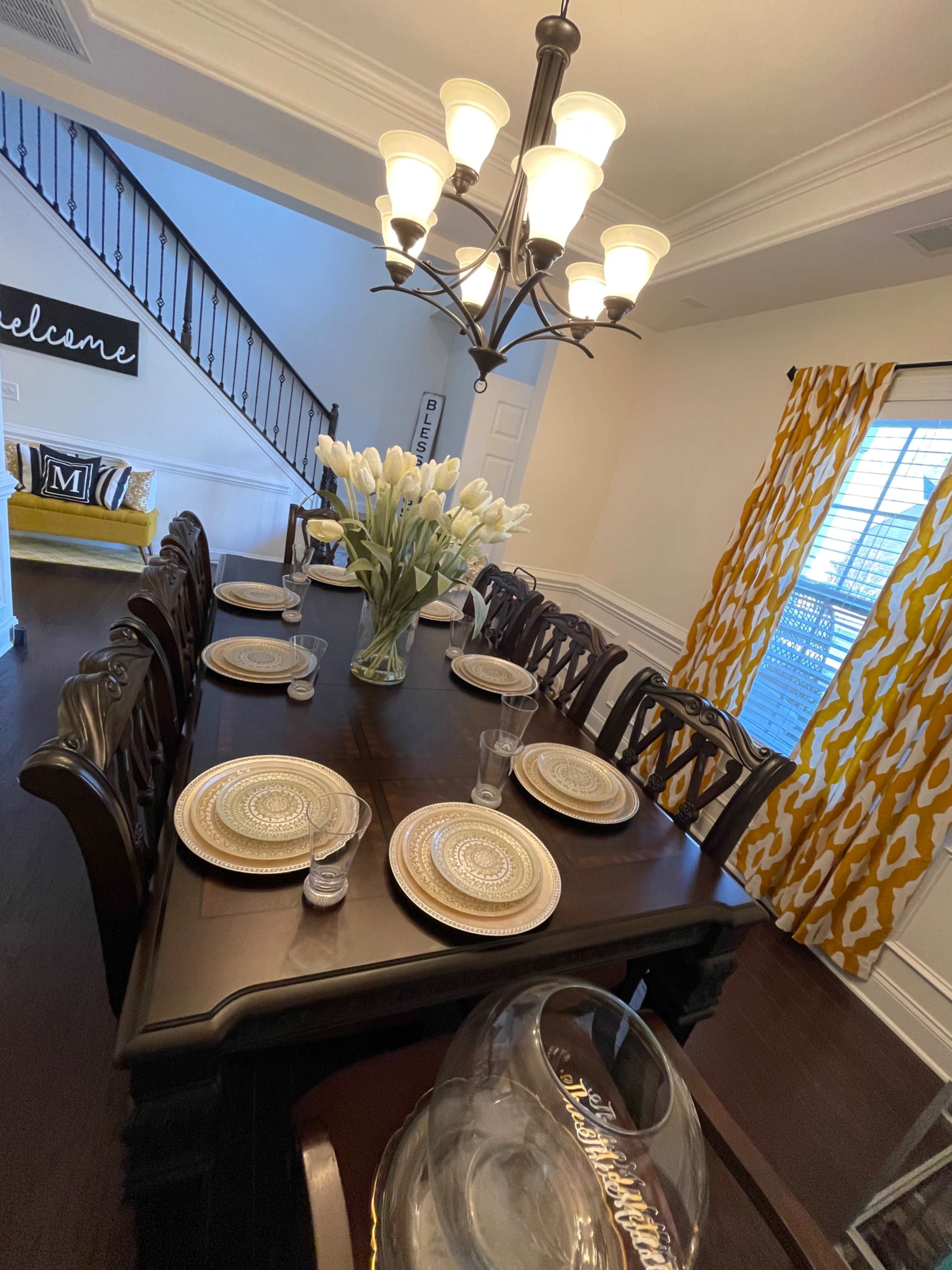 A spacious dining room features a long wooden table set with plates and glasses, surrounded by ornate chairs, with a chandelier above and a vase of tulips as a centerpiece.