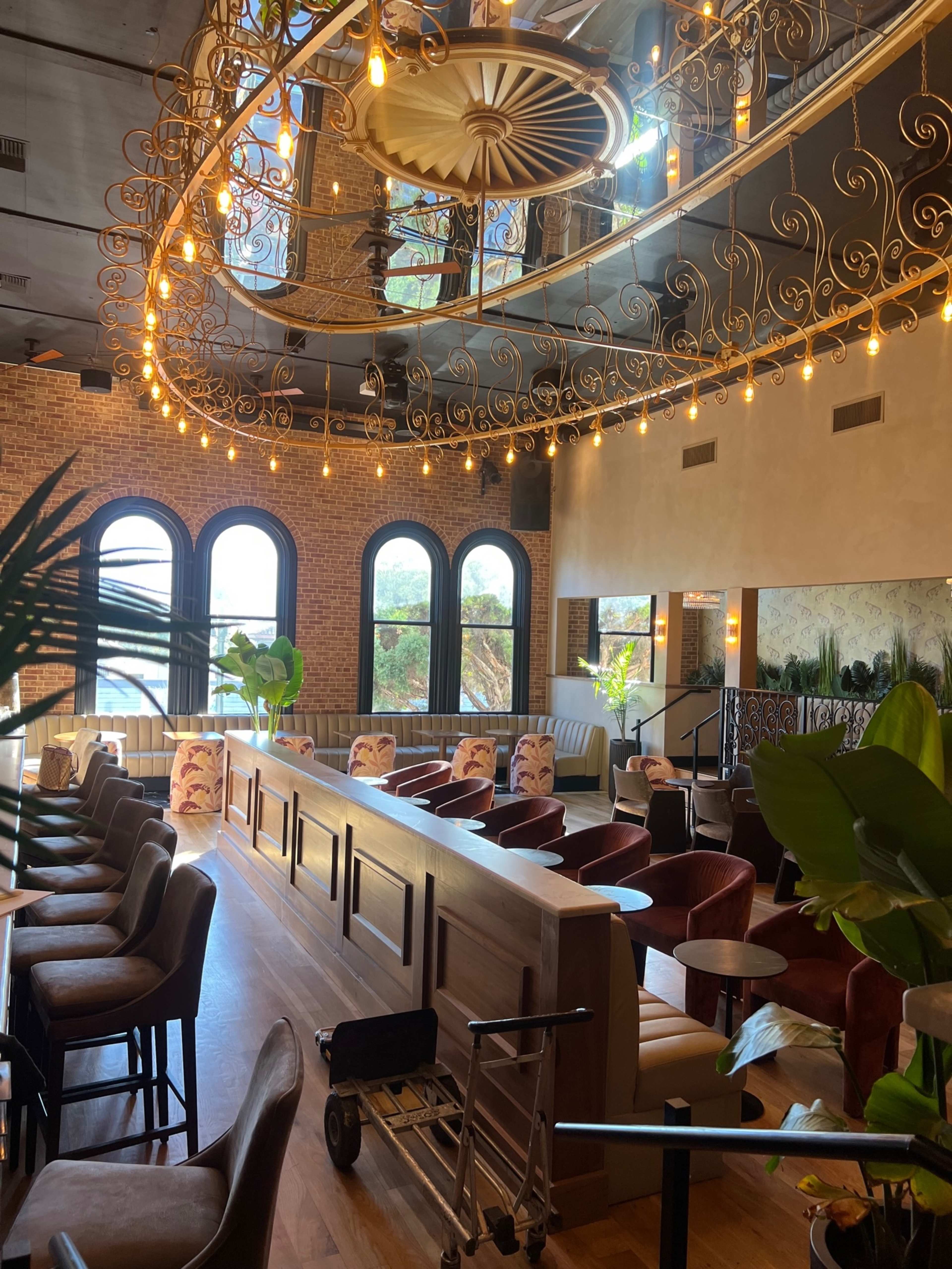 The image shows an upscale restaurant interior featuring a large, decorative chandelier, brick walls, and a mix of seating arrangements, including tables and chairs.