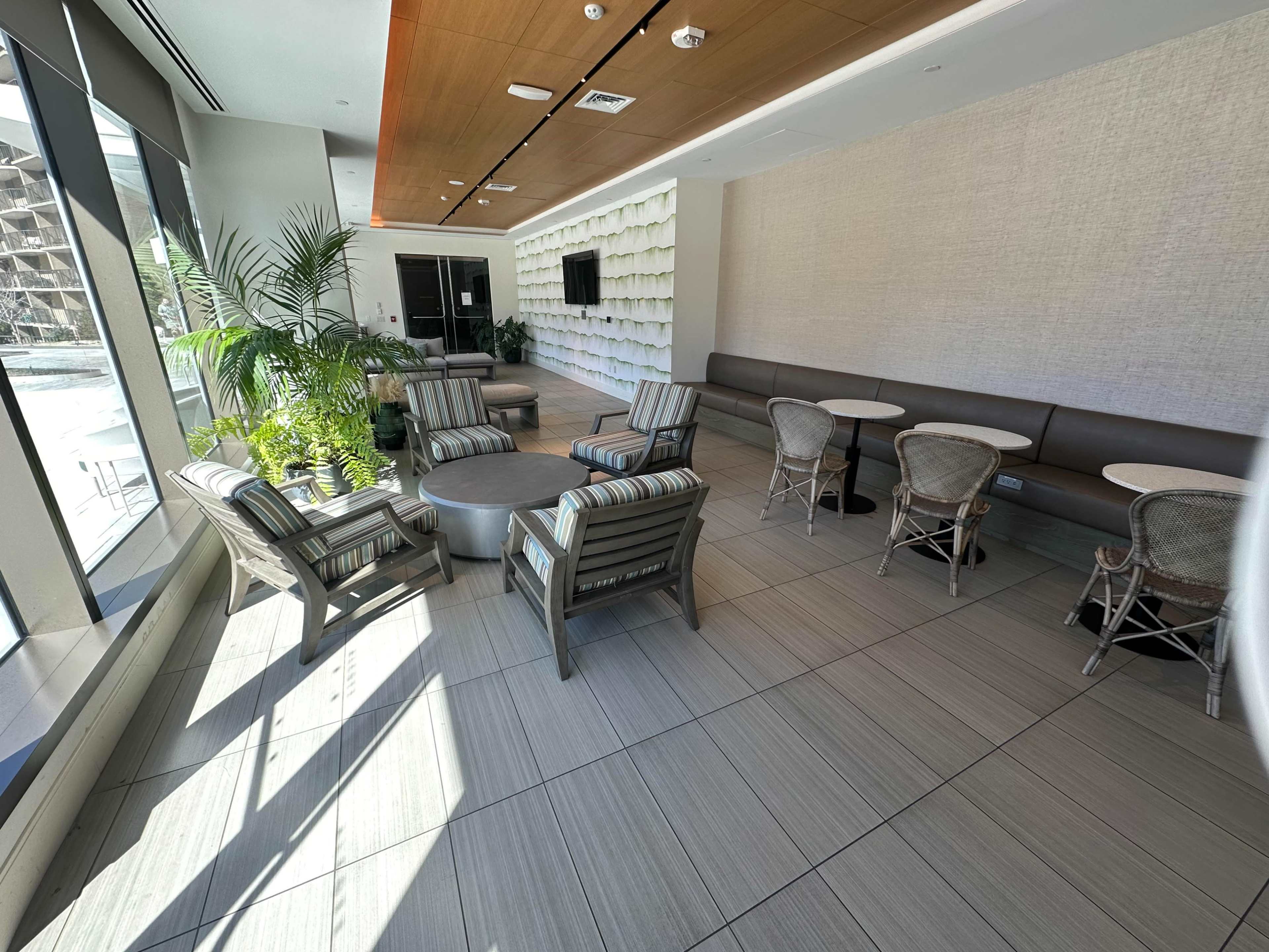 A modern lounge area features seating arrangements with striped chairs and a round table, surrounded by potted plants and large windows.
