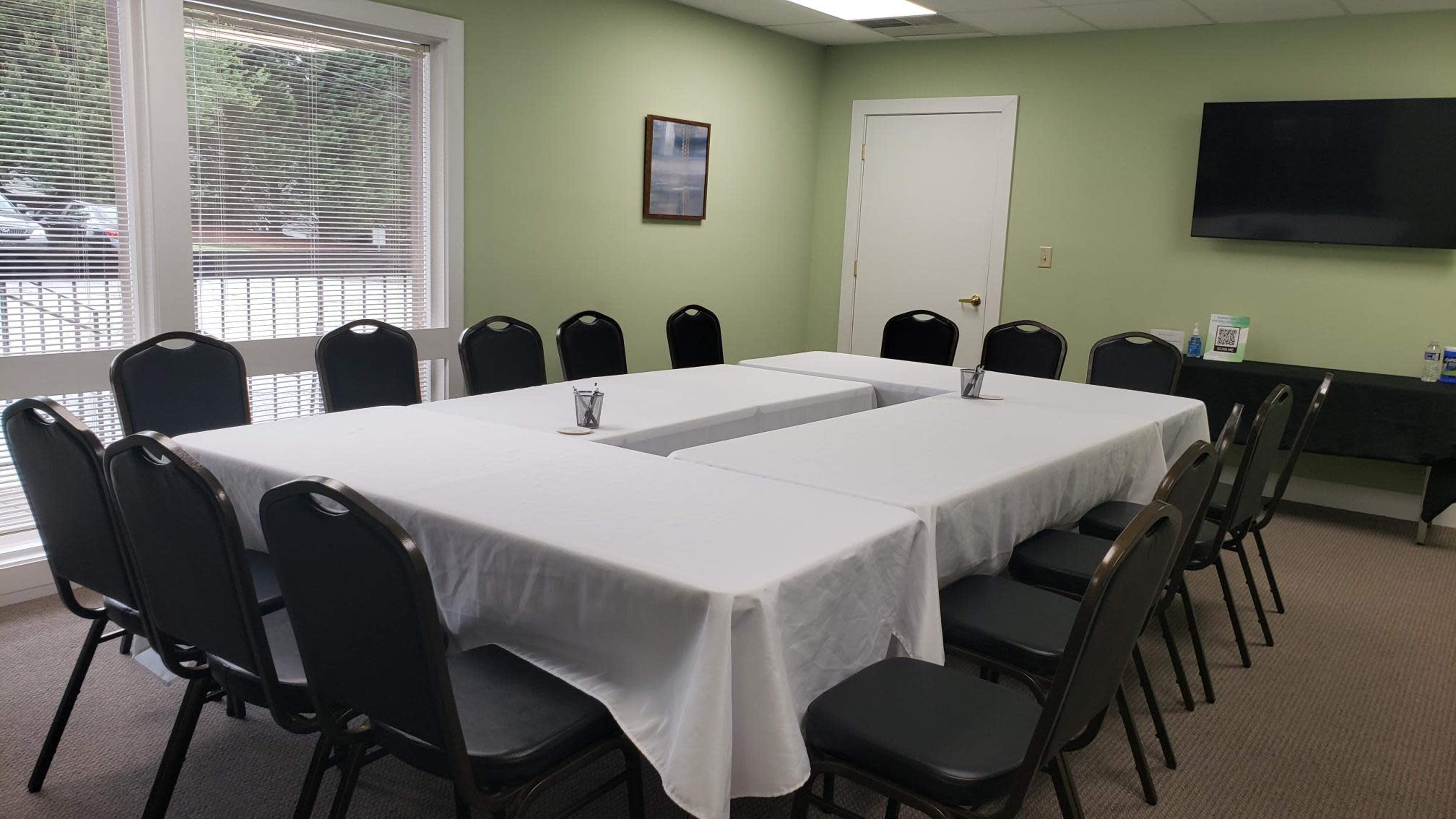 A meeting room features several rectangular tables arranged in a U-shape, covered with white tablecloths, surrounded by black chairs, and includes a television on the wall.