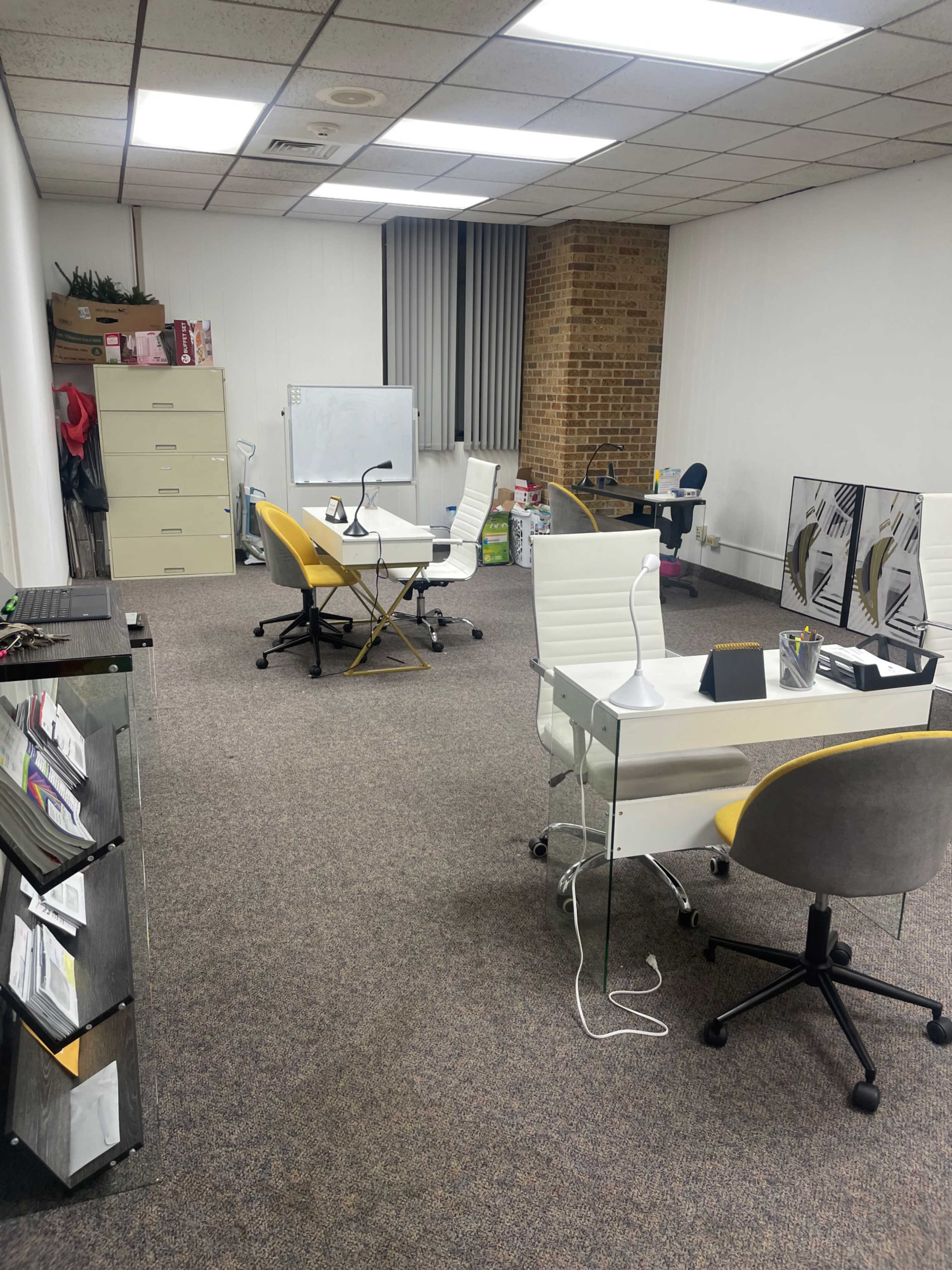 A sparsely furnished office space with two desks, a filing cabinet, and a whiteboard against the wall.