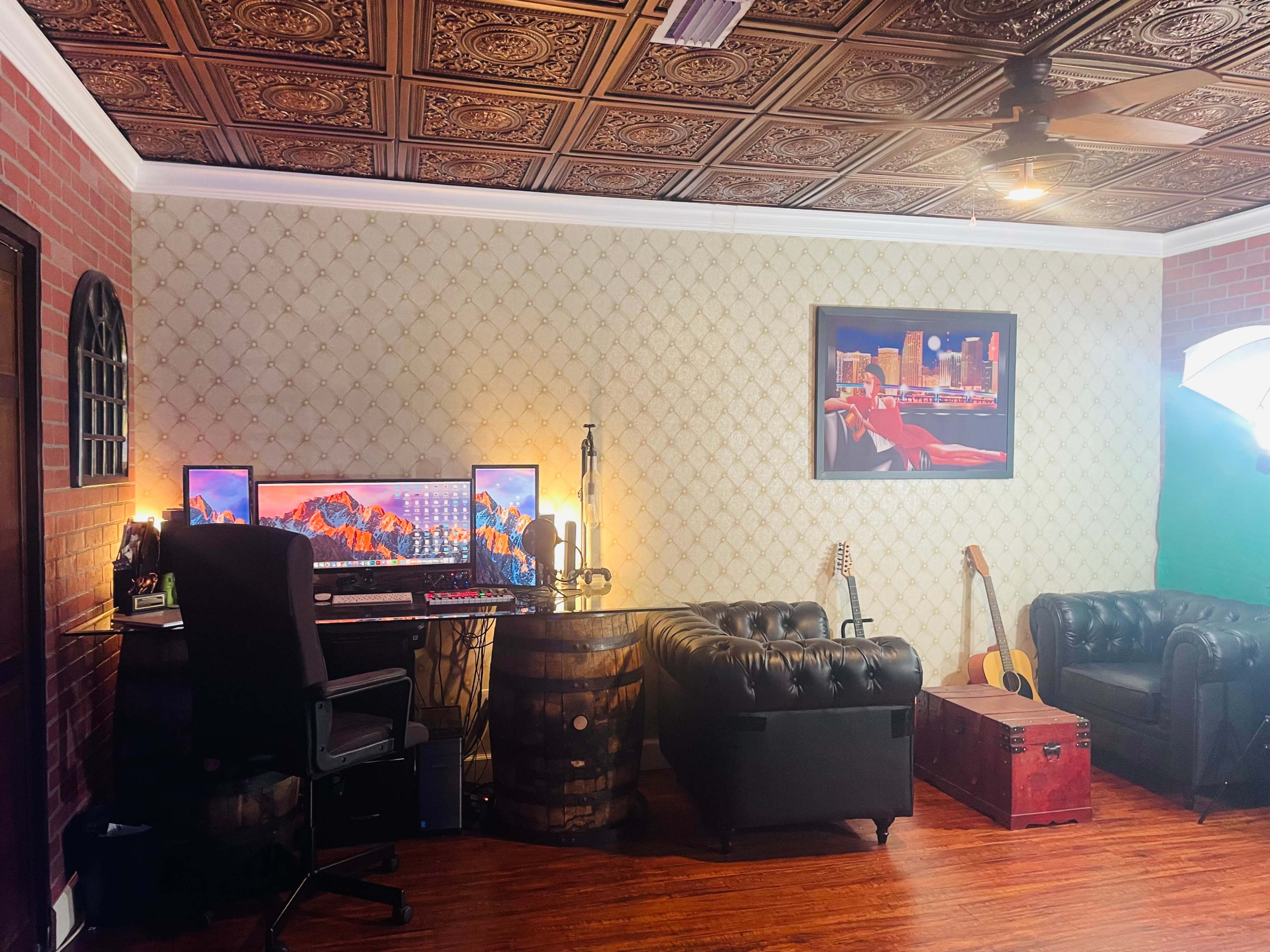 The image shows a stylish office space with a computer setup, two leather couches, a guitar, and decorative wall art.