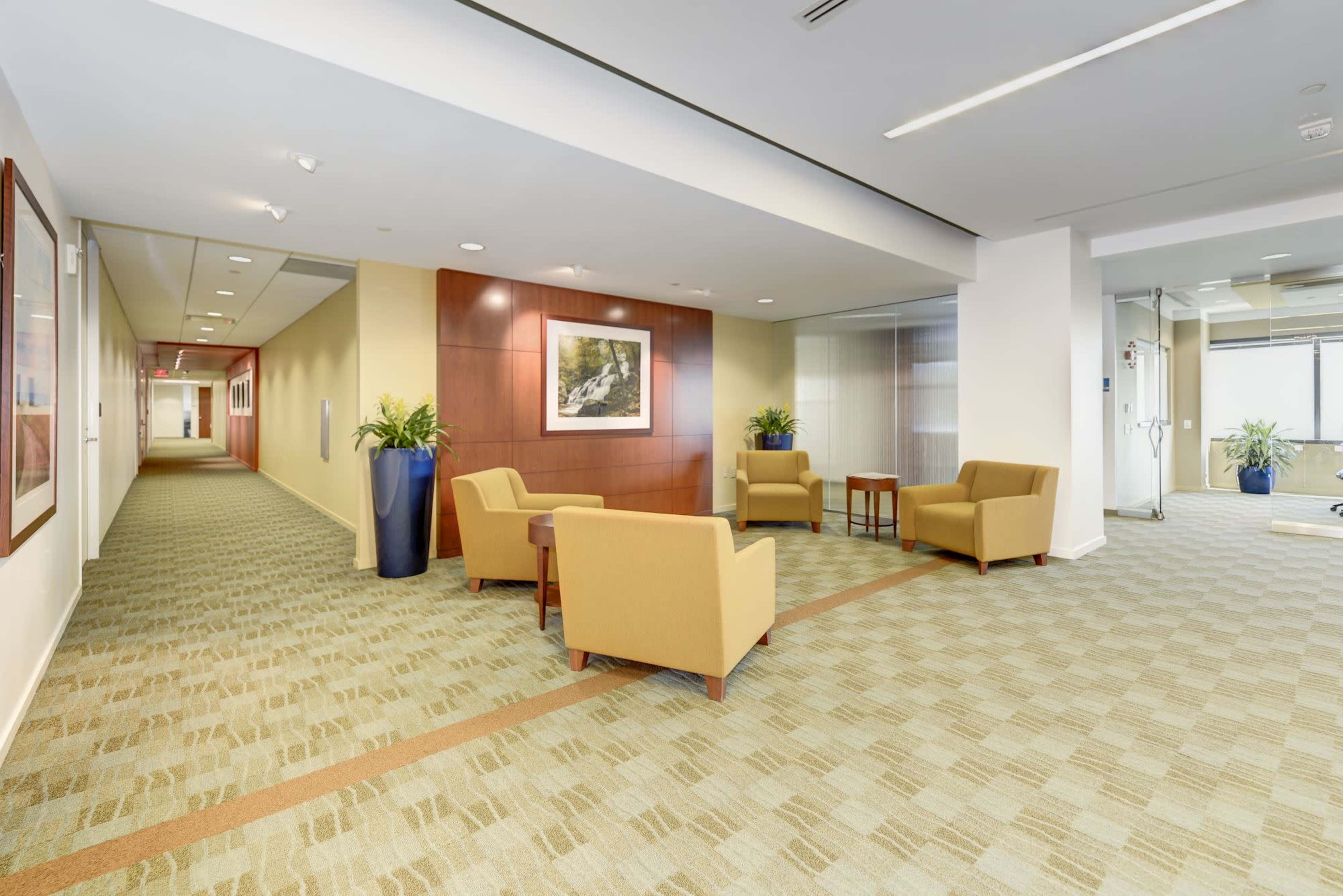 A modern office corridor features a seating area with two chairs and a small table, alongside a decorated wall and a well-lit hallway.