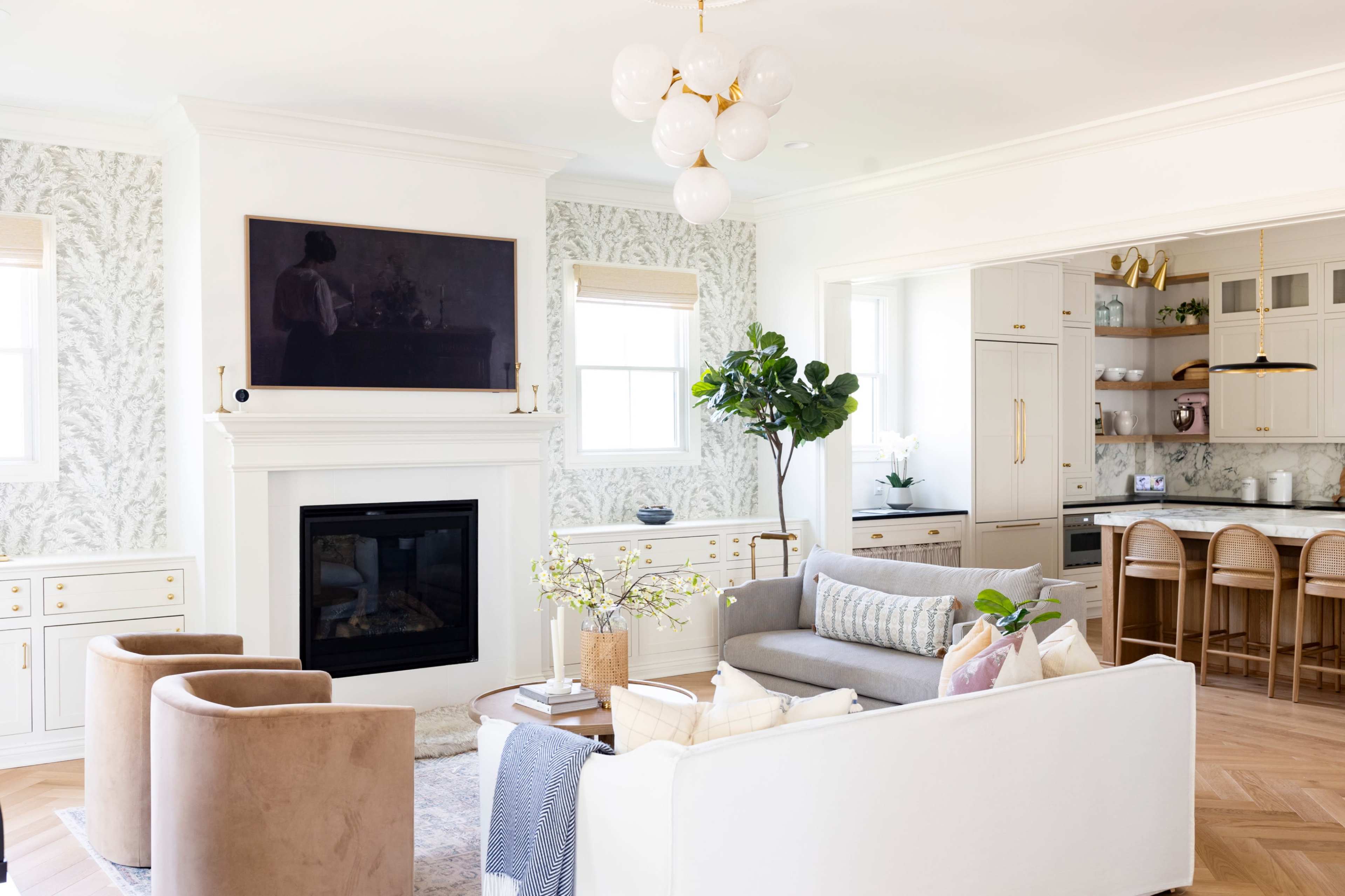 The image depicts a modern living room featuring a white sofa, two beige chairs, a fireplace with a large mounted television, and a kitchen area in the background with beige cabinetry and bar stools.