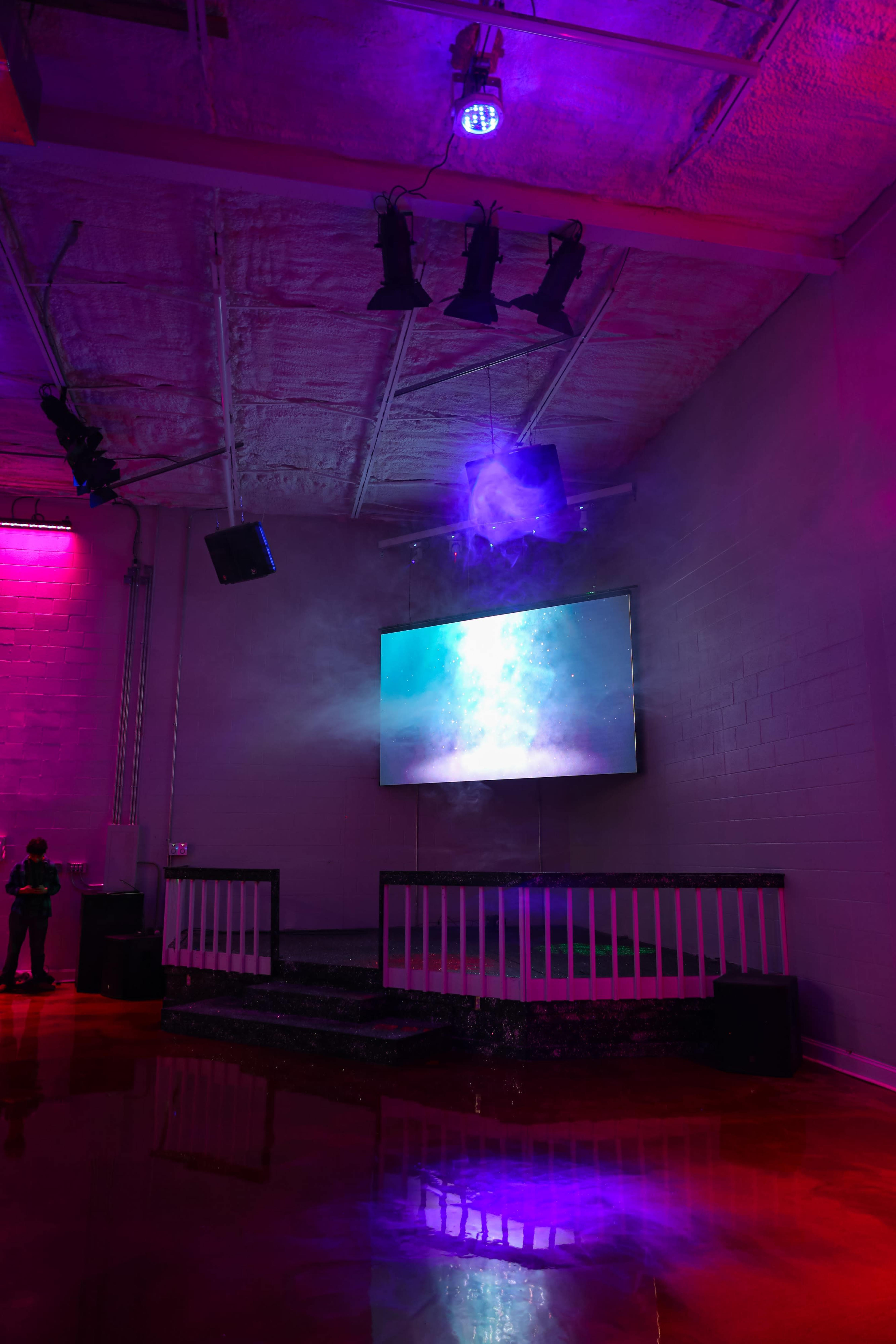 A dimly lit venue features a stage with a large screen displaying a colorful visual, surrounded by purple lighting and smoke effects.