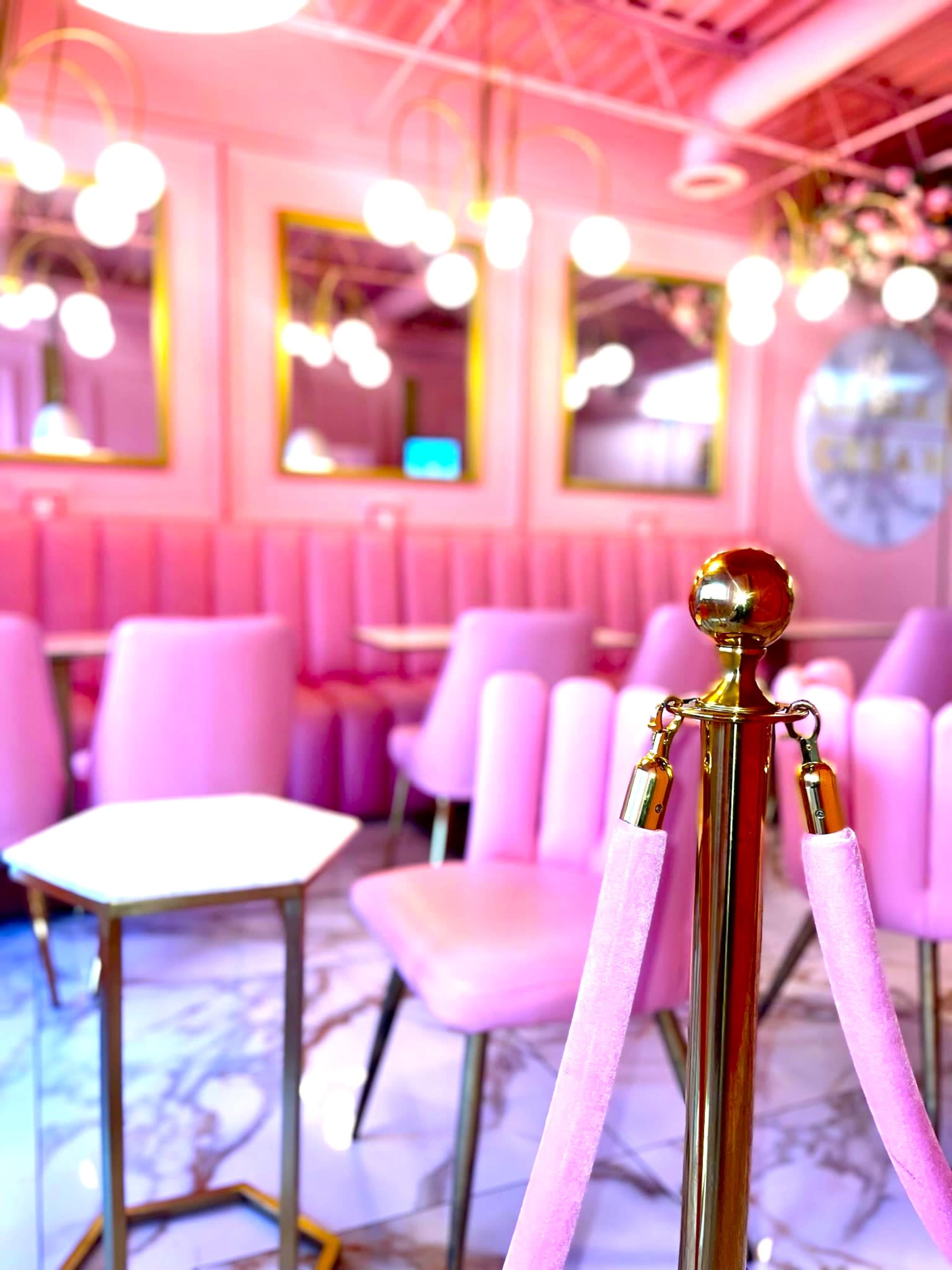 A pink-themed café interior featuring plush seating, decorative mirrors, and round pendant lights.