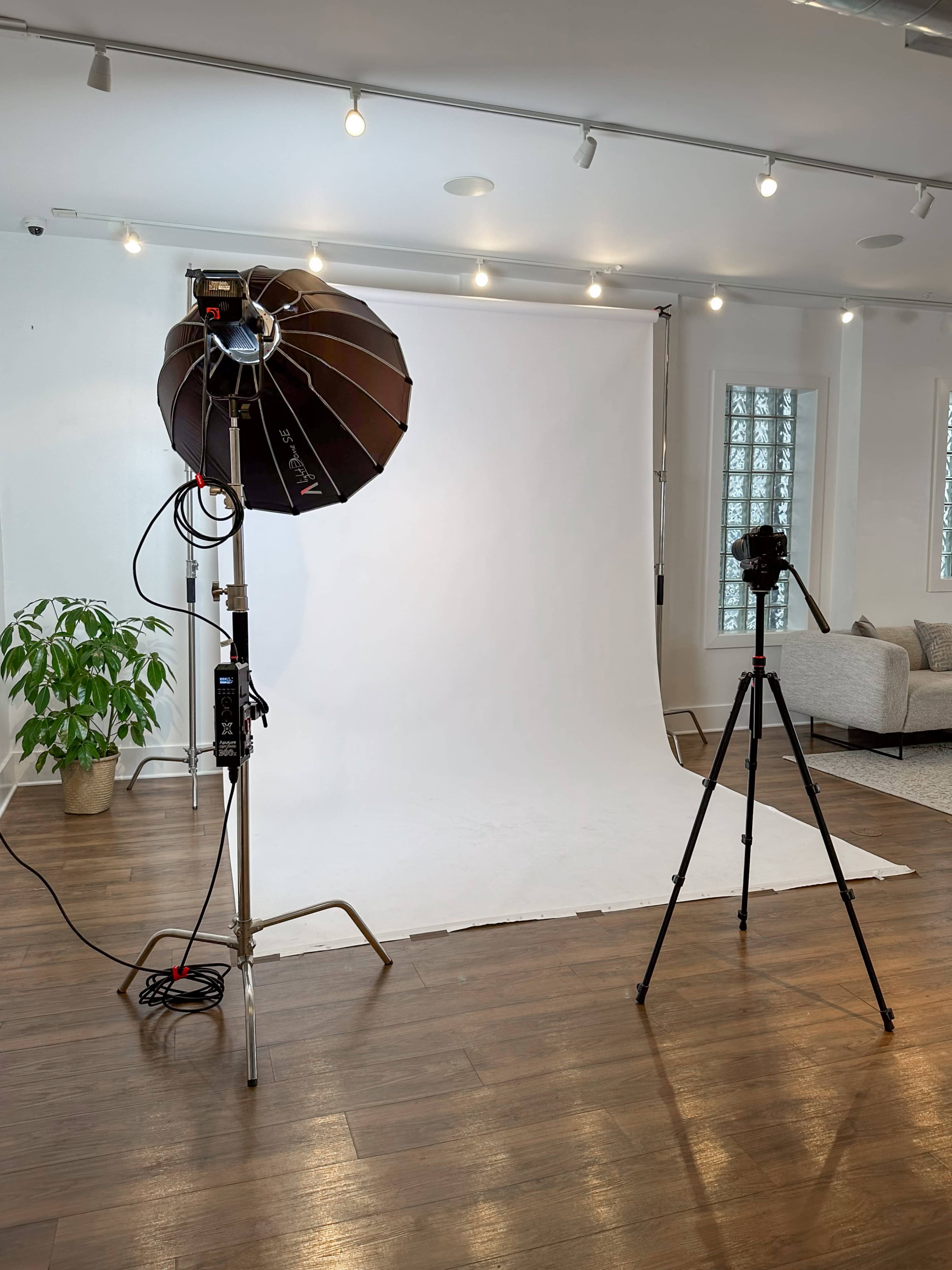 A photography studio is set up with a softbox light, a camera on a tripod, and a plain white backdrop.