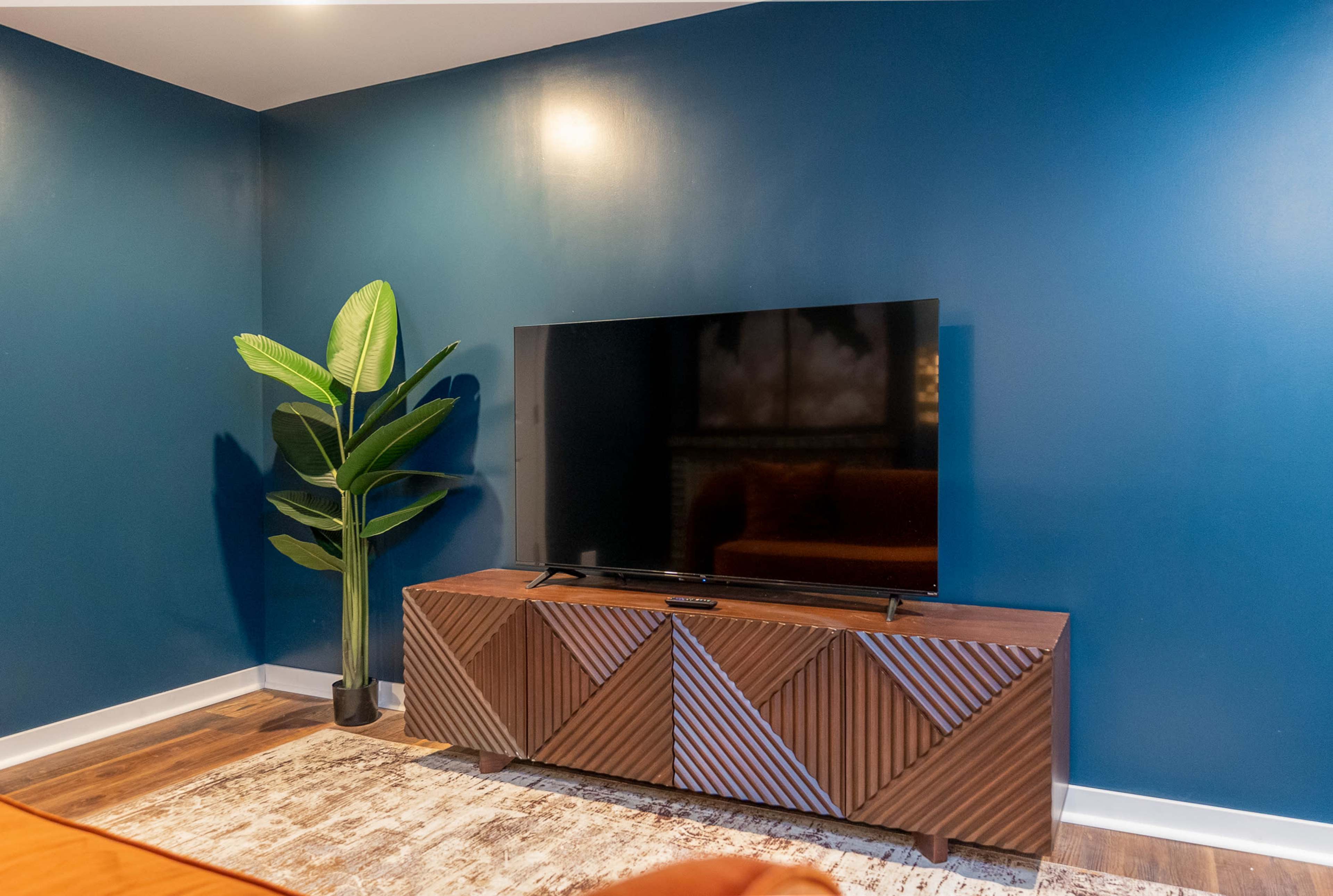 A modern living room corner with a television mounted on a wooden cabinet in front of a dark blue wall, accompanied by a tall green plant.