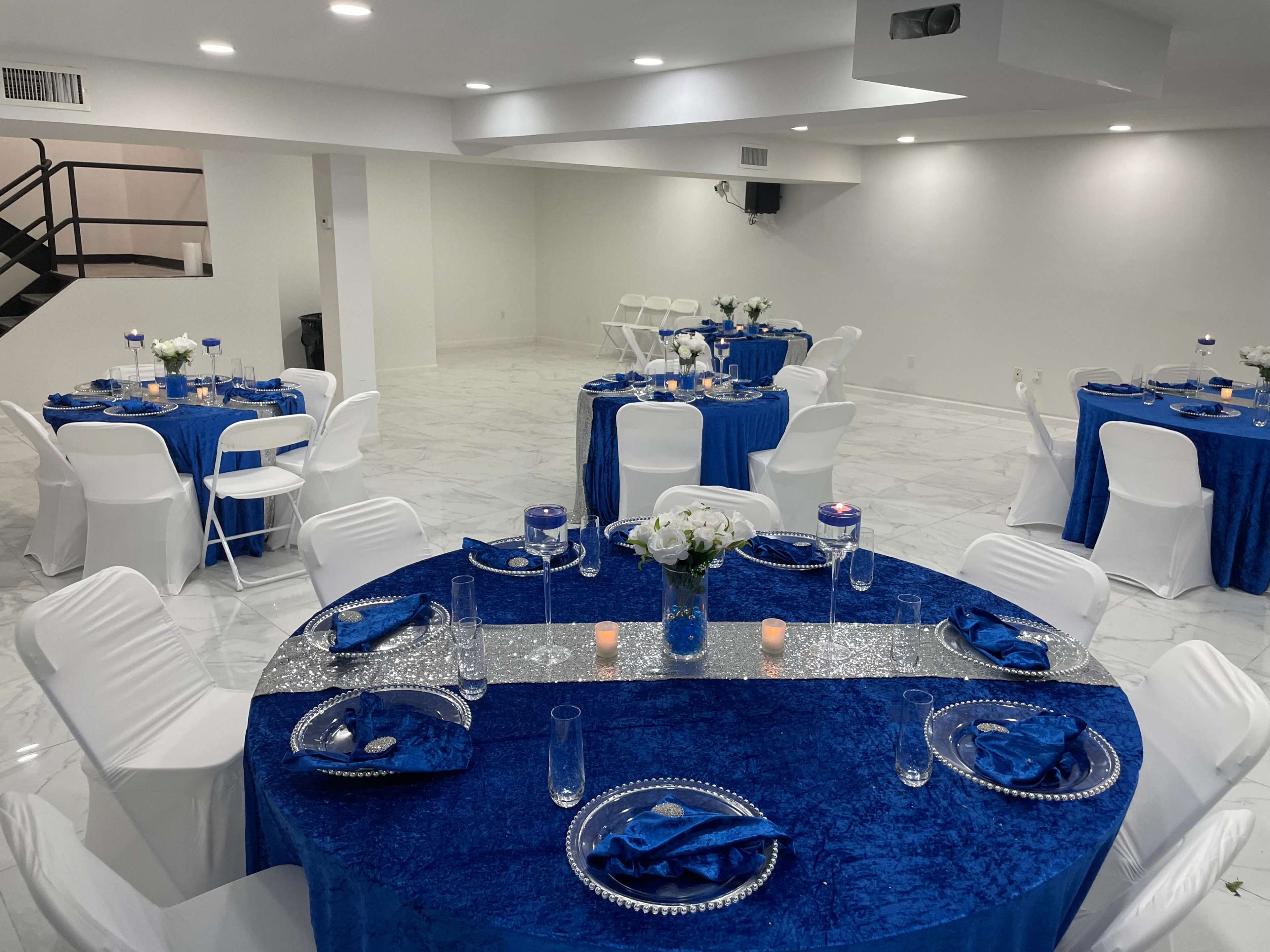 A spacious event room with white walls and marble flooring, featuring round tables covered in blue tablecloths, each set with white chairs and decorative centerpieces.