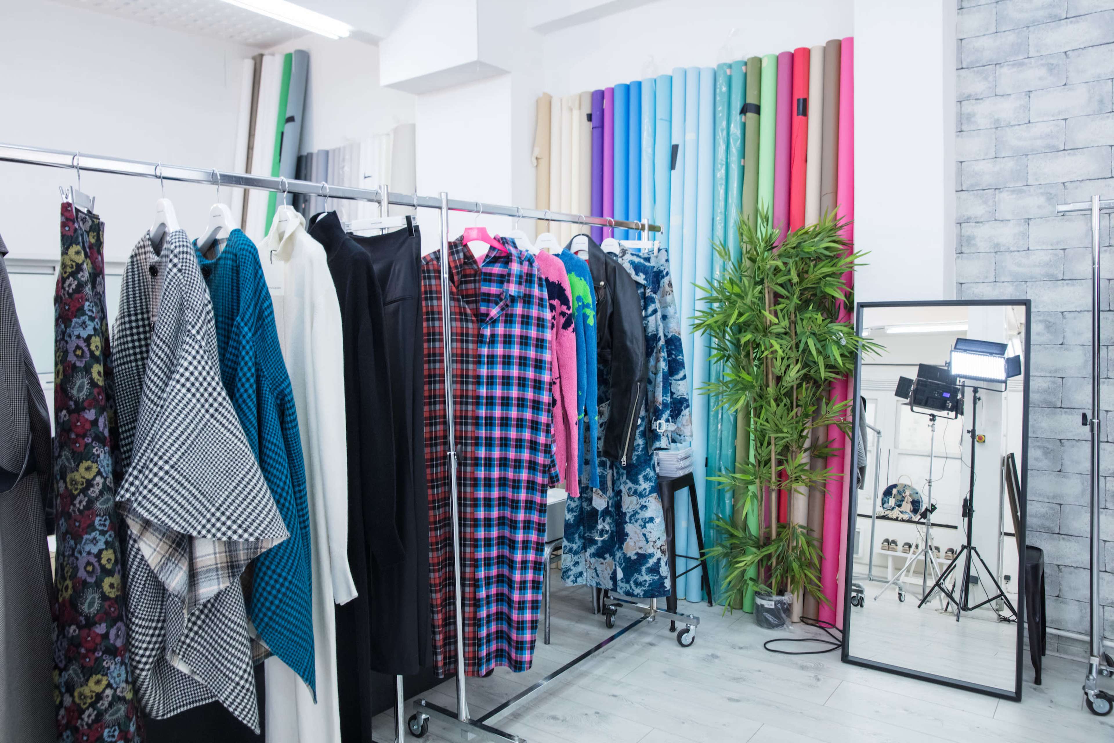 The image shows a clothing rack filled with various garments in a brightly lit fashion studio, with colorful backdrops and a mirror reflecting the space.