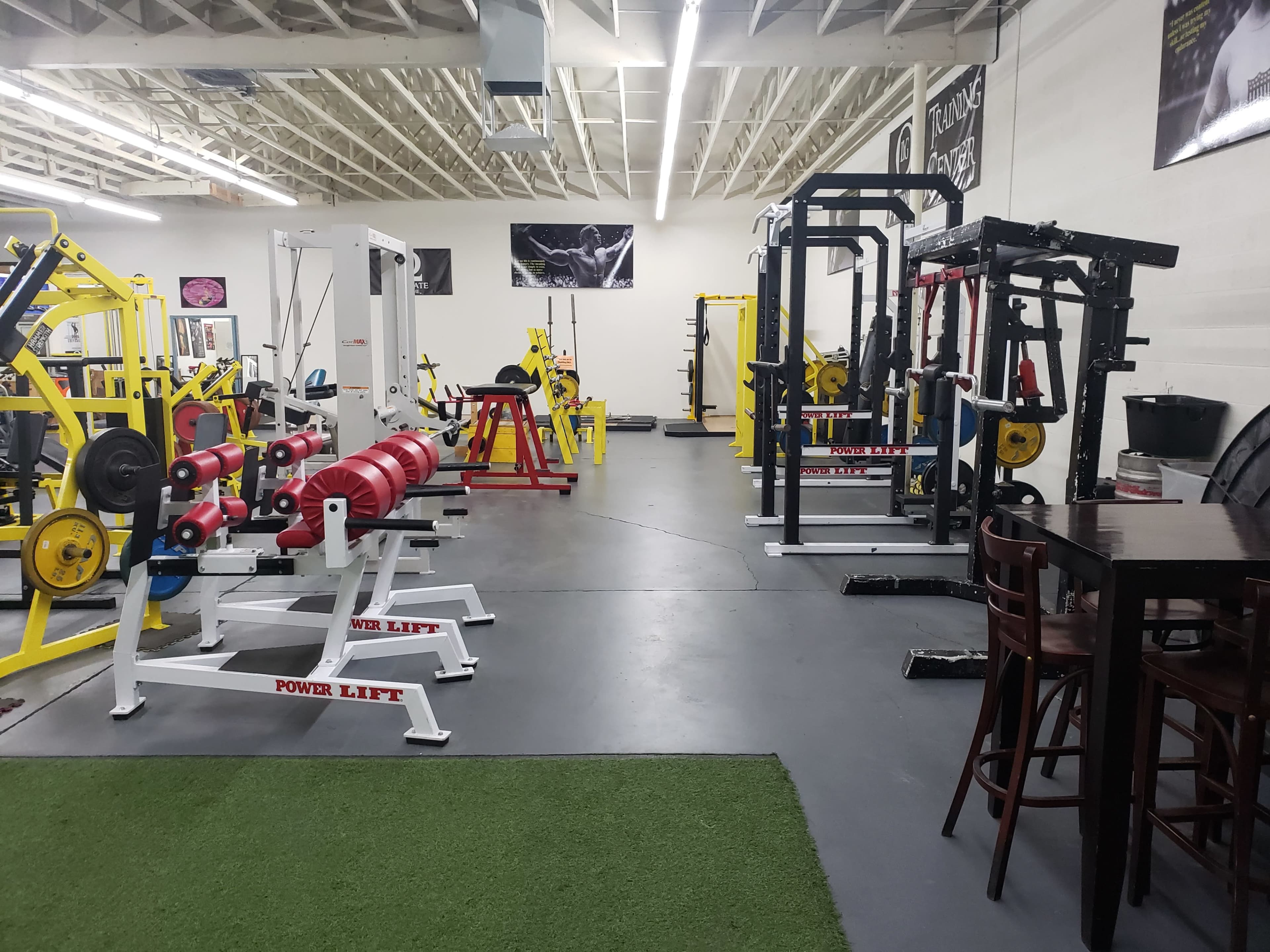 The image shows a spacious gym with various weightlifting equipment, including benches, squat racks, and free weights, arranged in an organized layout.