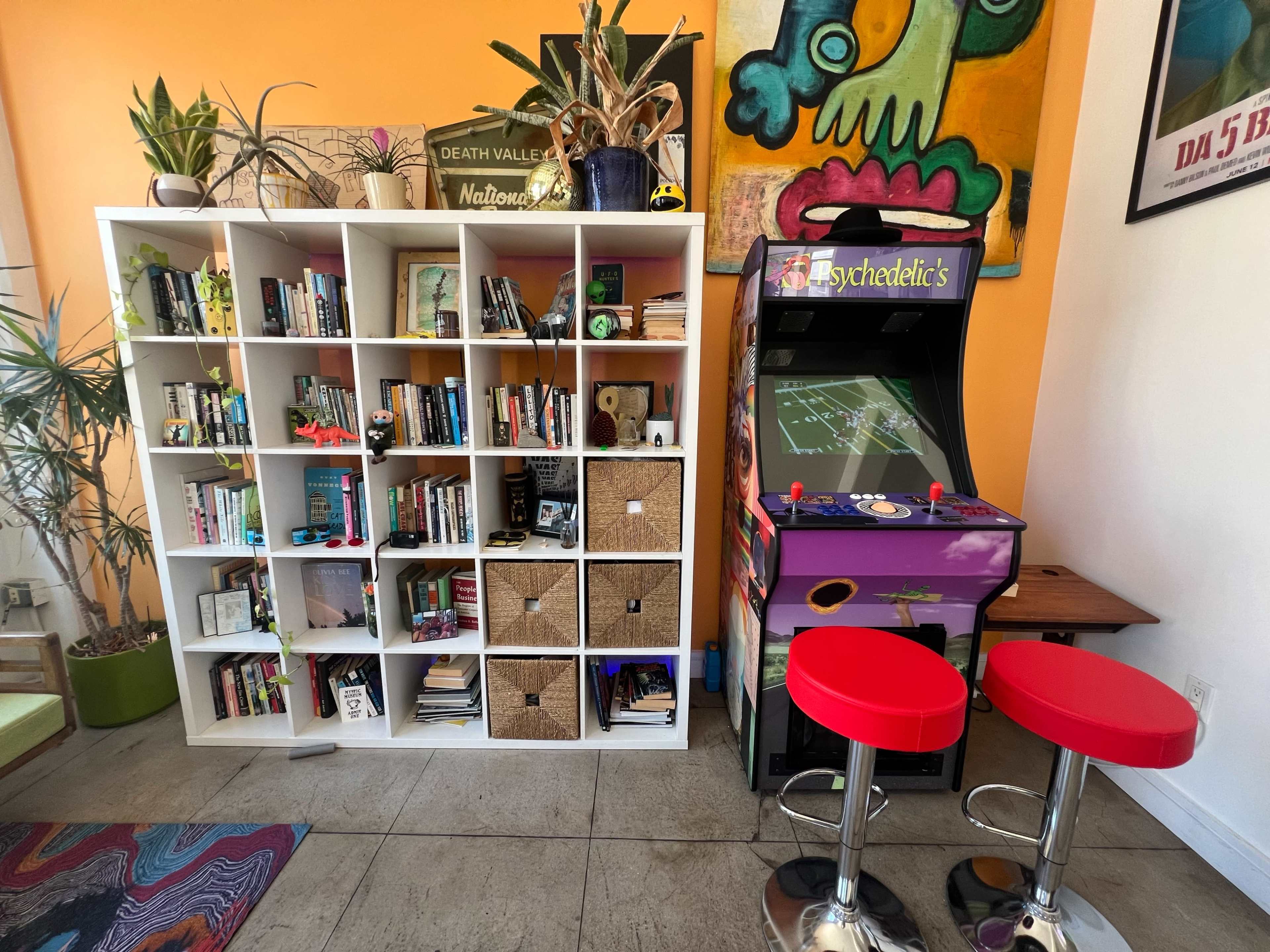 The scene features a white bookshelf filled with books and decorative items beside a vibrant arcade game cabinet, with red bar stools in front.