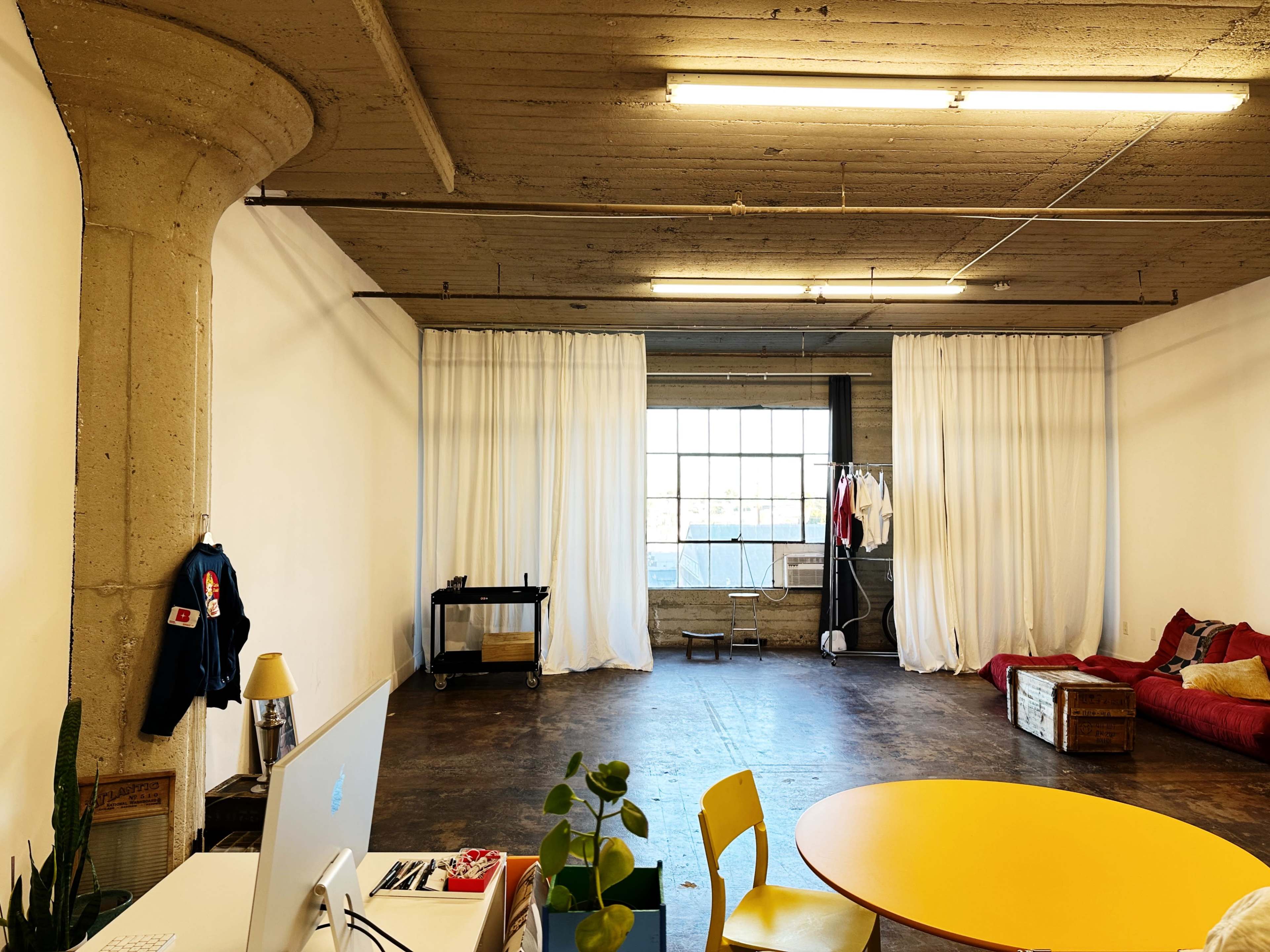 The image shows a spacious room with a large window covered by sheer curtains, a yellow round table, a red couch, and a workspace with a computer.