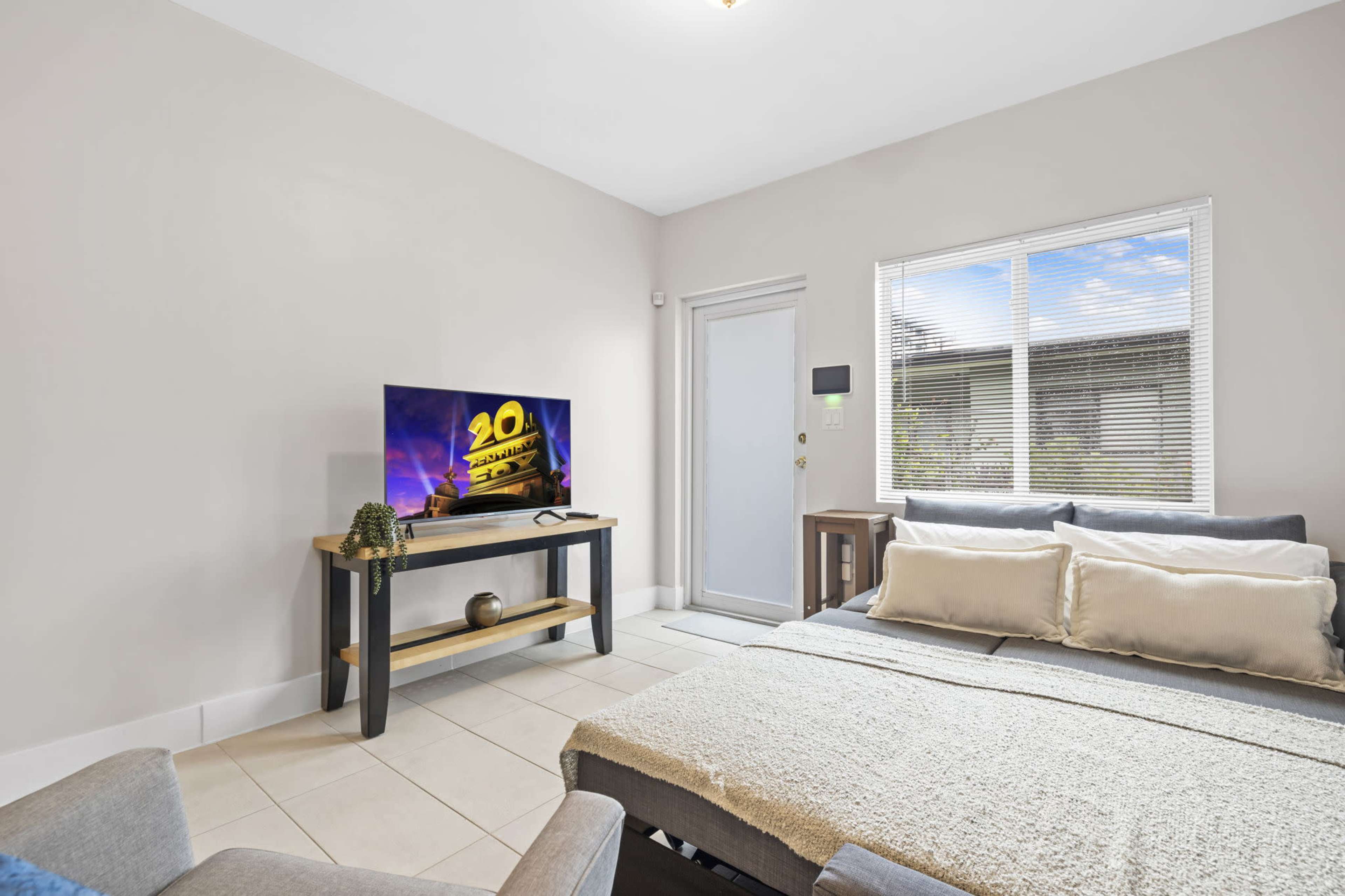 The image shows a small living area with a television on a wooden stand, a sofa, and a door leading outside, all set against a light gray wall.