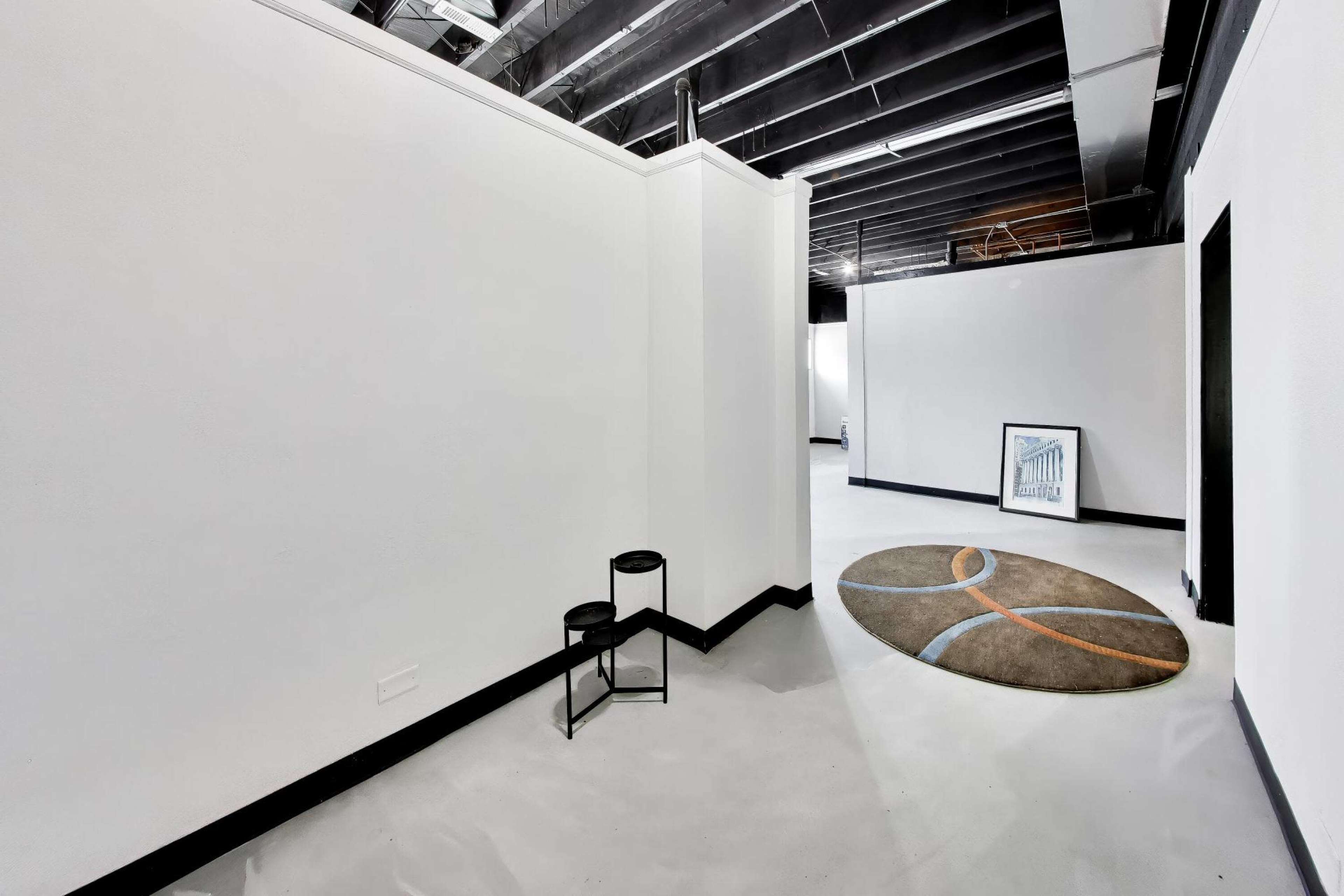 The image depicts a spacious, minimalist interior with white walls, concrete flooring, and a round rug featuring a simple design.