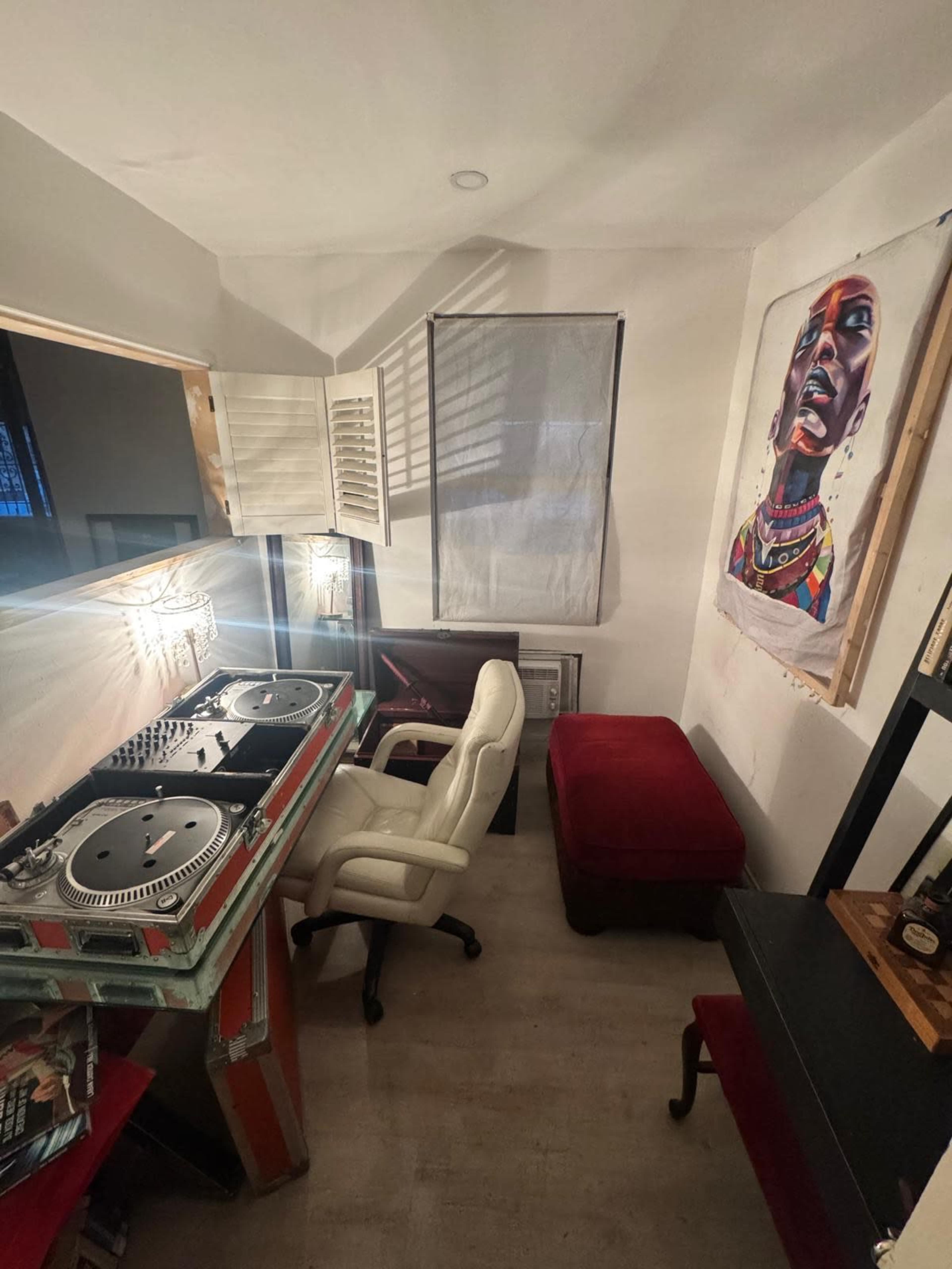 The image shows a small room with a DJ setup featuring turntables on a table, a white chair, a red ottoman, and a colorful artwork on the wall.