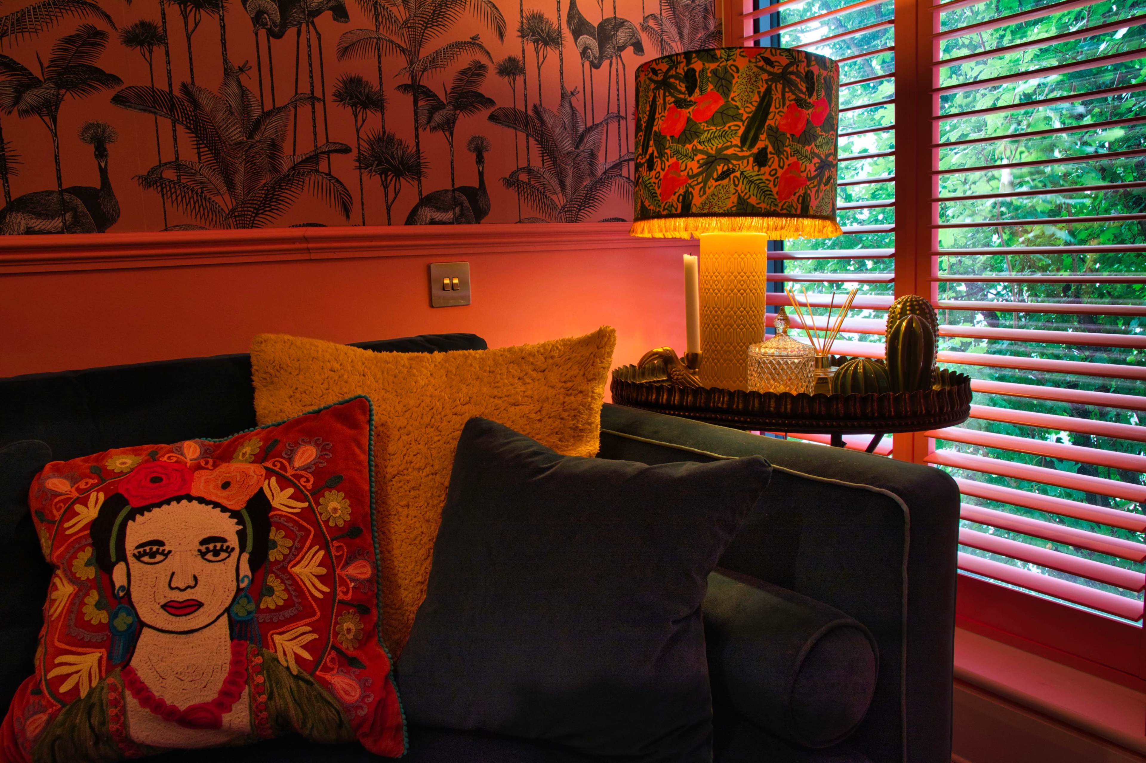 A brightly decorated living room features a blue couch with colorful pillows, a floral lamp on a side table, and plant-patterned wallpaper.