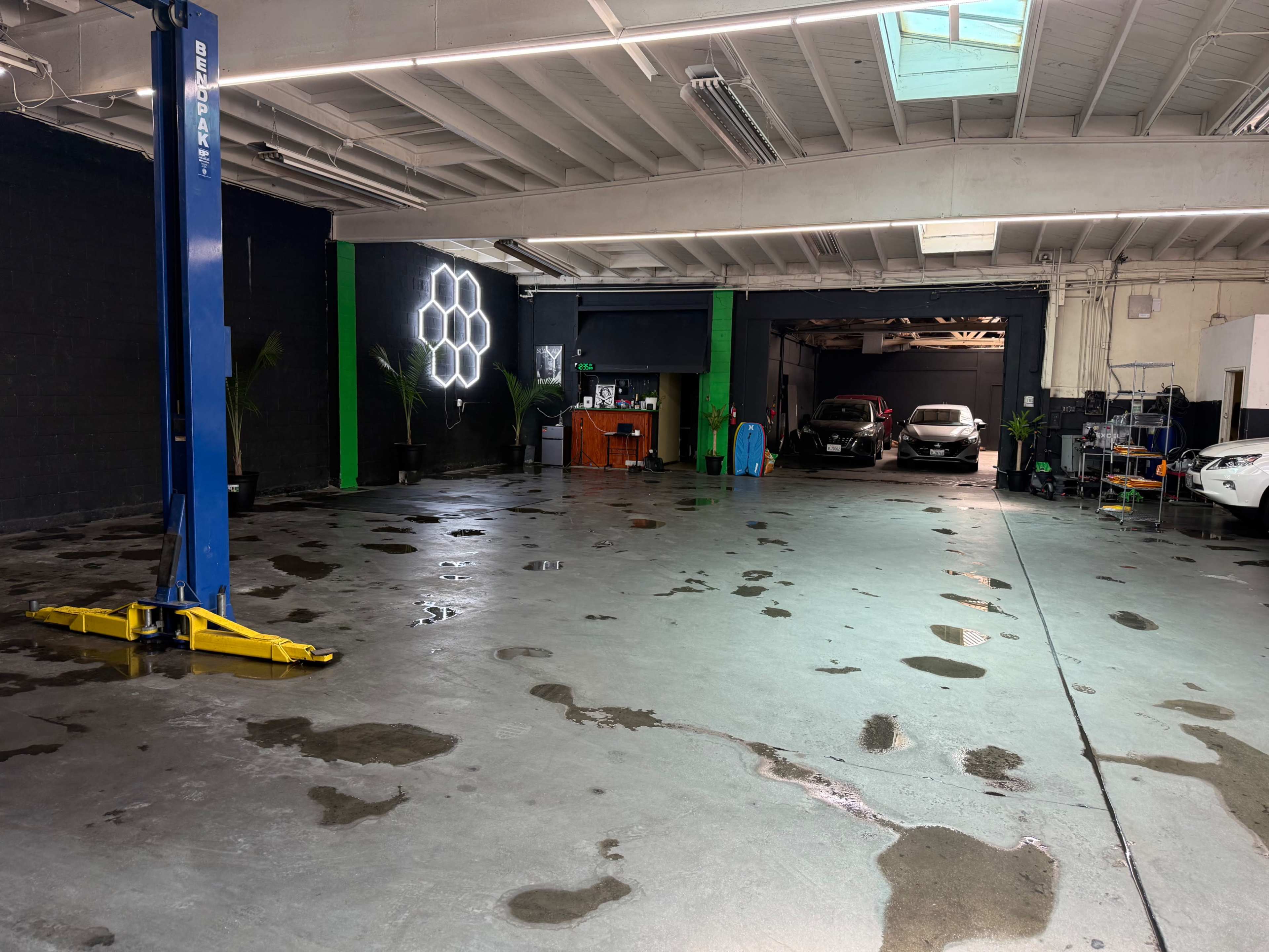 The image shows a spacious garage with a concrete floor, featuring a blue car lift on the left and several vehicles parked in the background, alongside bright green and black walls.