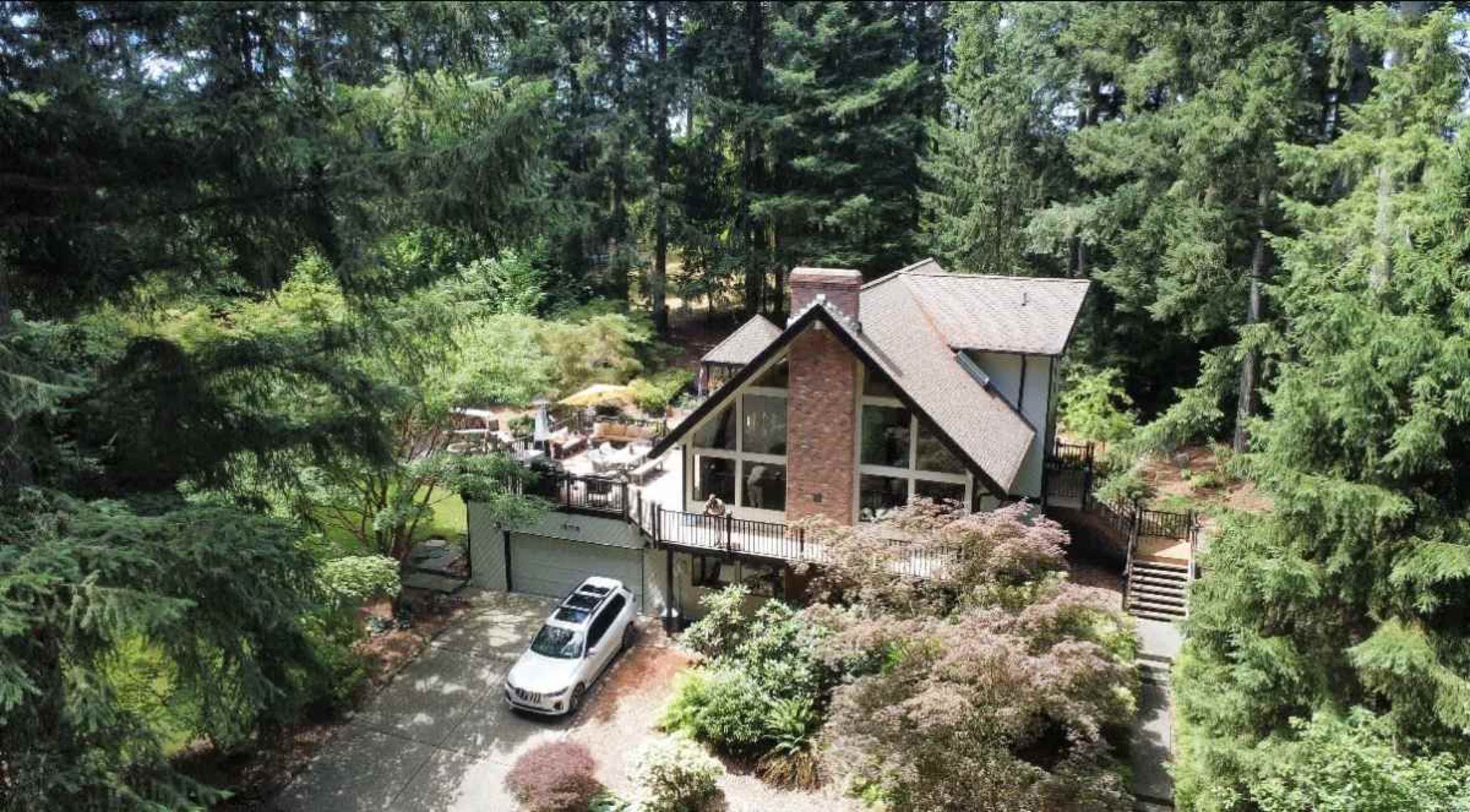A large, modern house with a sloping roof is surrounded by tall trees and lush greenery, featuring a driveway and an outdoor patio area.
