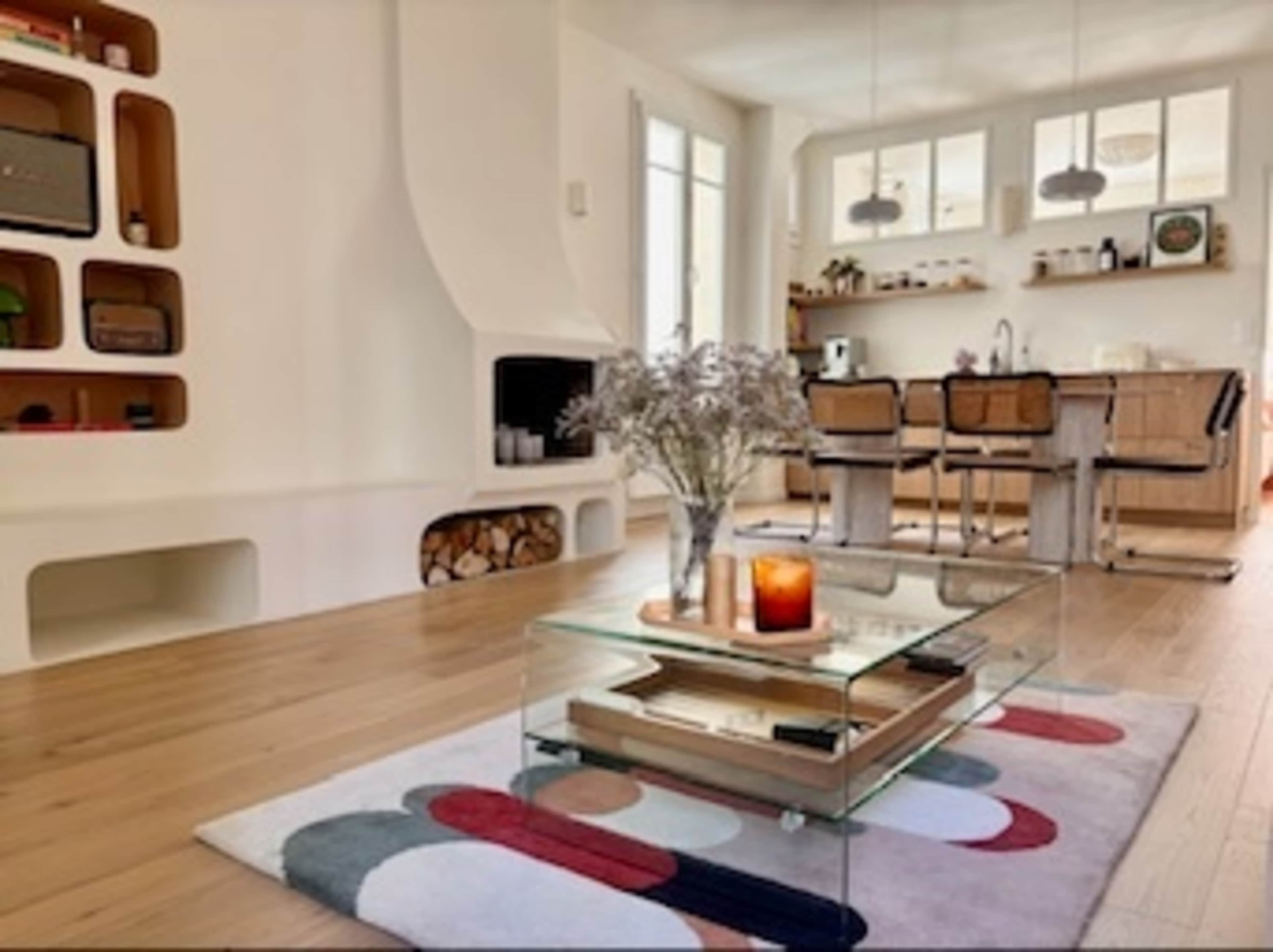The image shows a modern living room with a glass coffee table, a colorful area rug, and a kitchen area visible in the background.