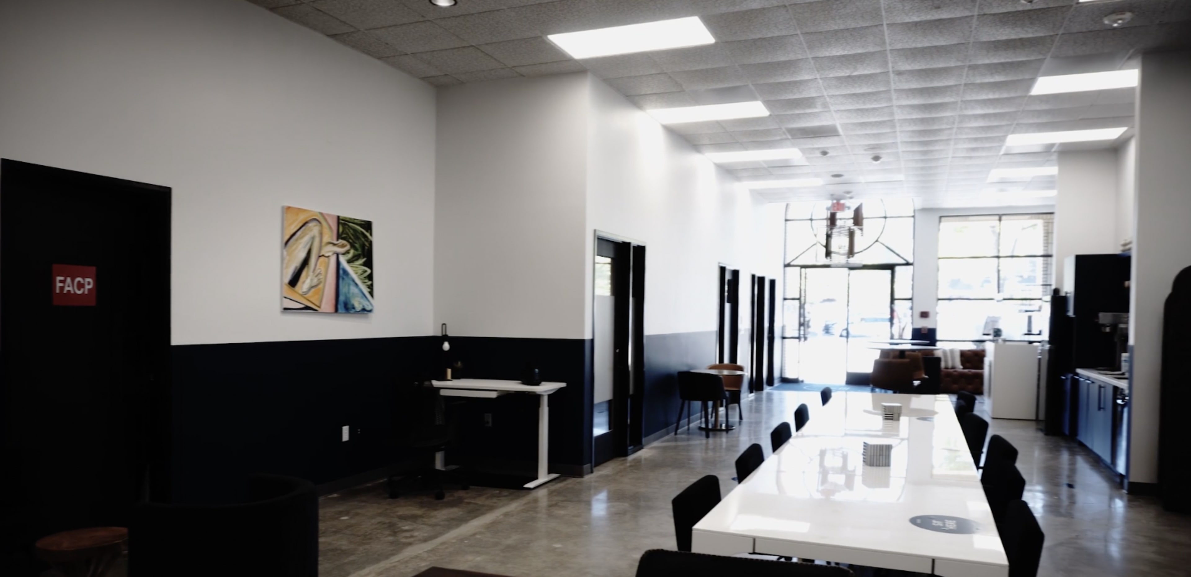 A hallway in a modern office features a long white table, black chairs, and windows at the far end, with artwork hanging on the wall.