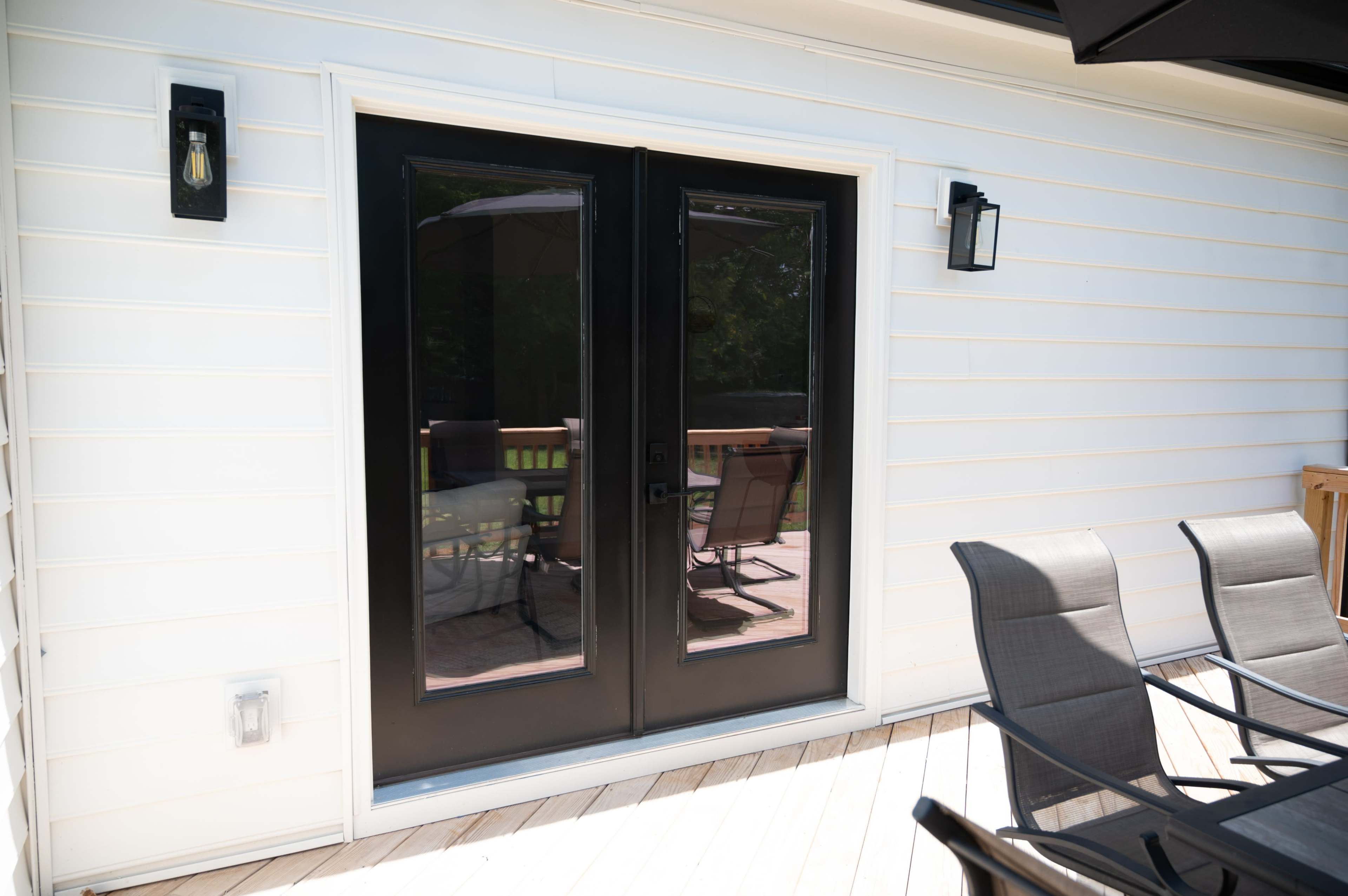 The image shows a set of black double doors leading to a deck, framed by white siding and flanked by outdoor lighting fixtures.