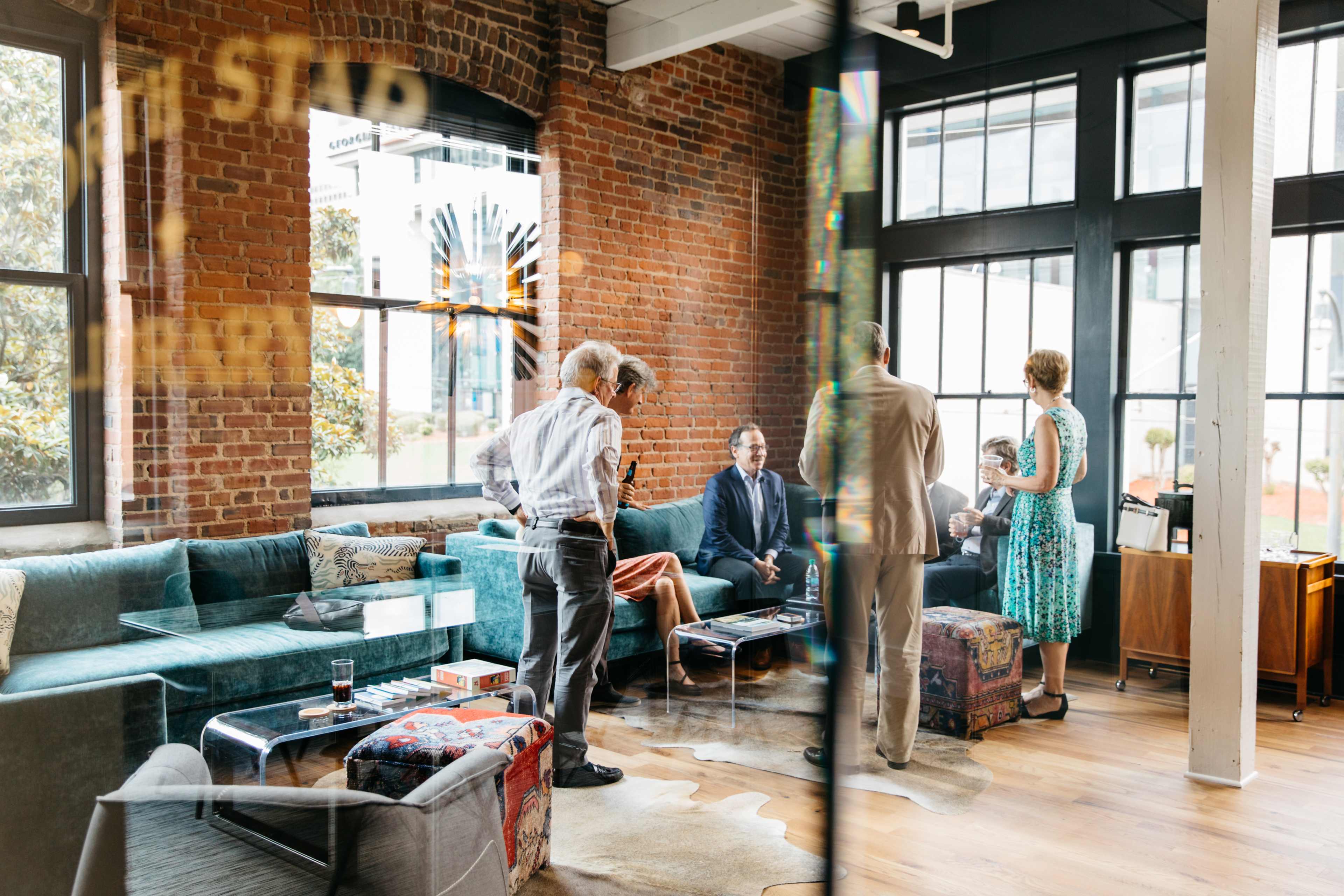A group of five people engages in conversation in a modern, stylish lounge with large windows and exposed brick walls.