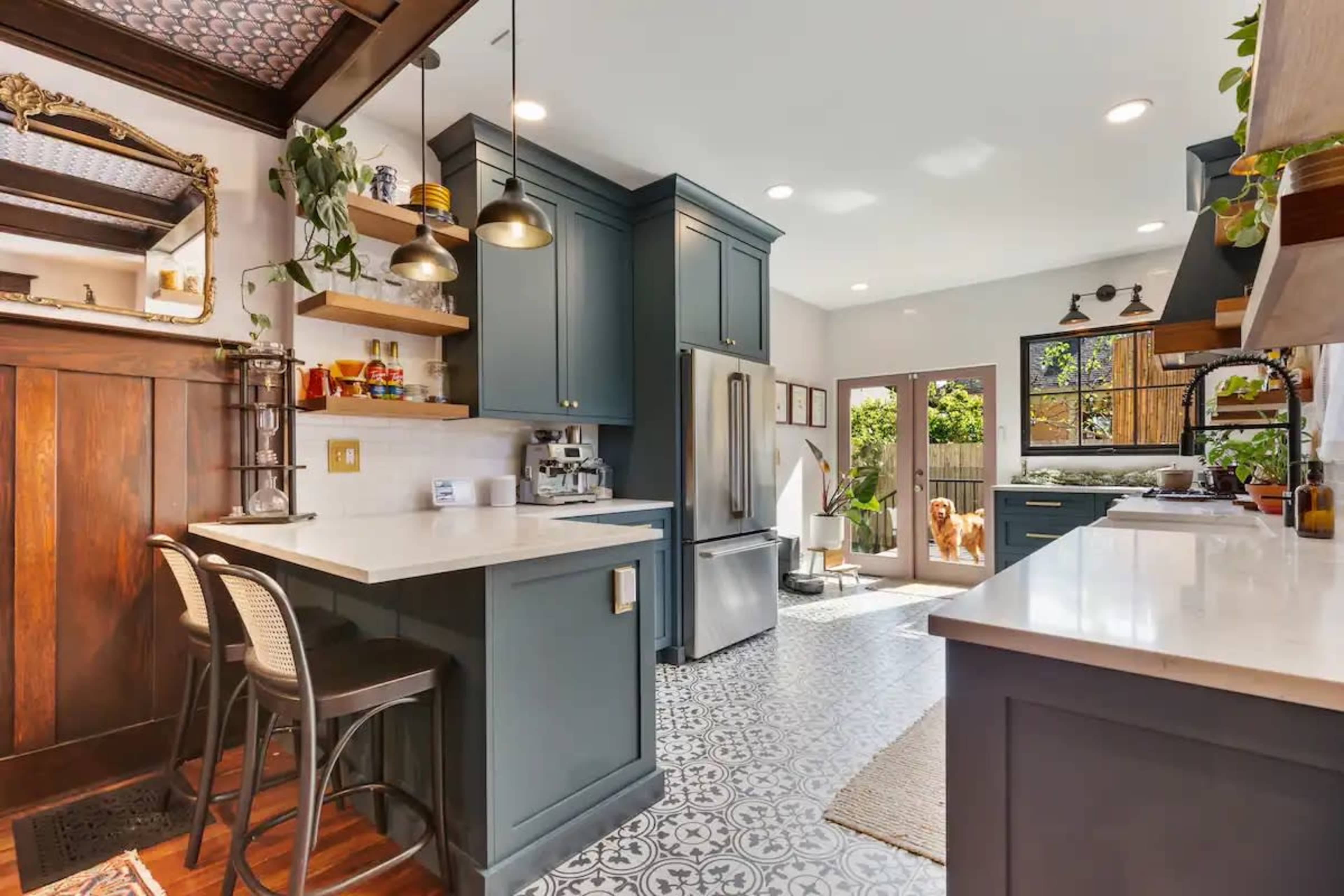 A modern kitchen features dark green cabinetry, white countertops, and patterned tile flooring, with an open door leading to a backyard.