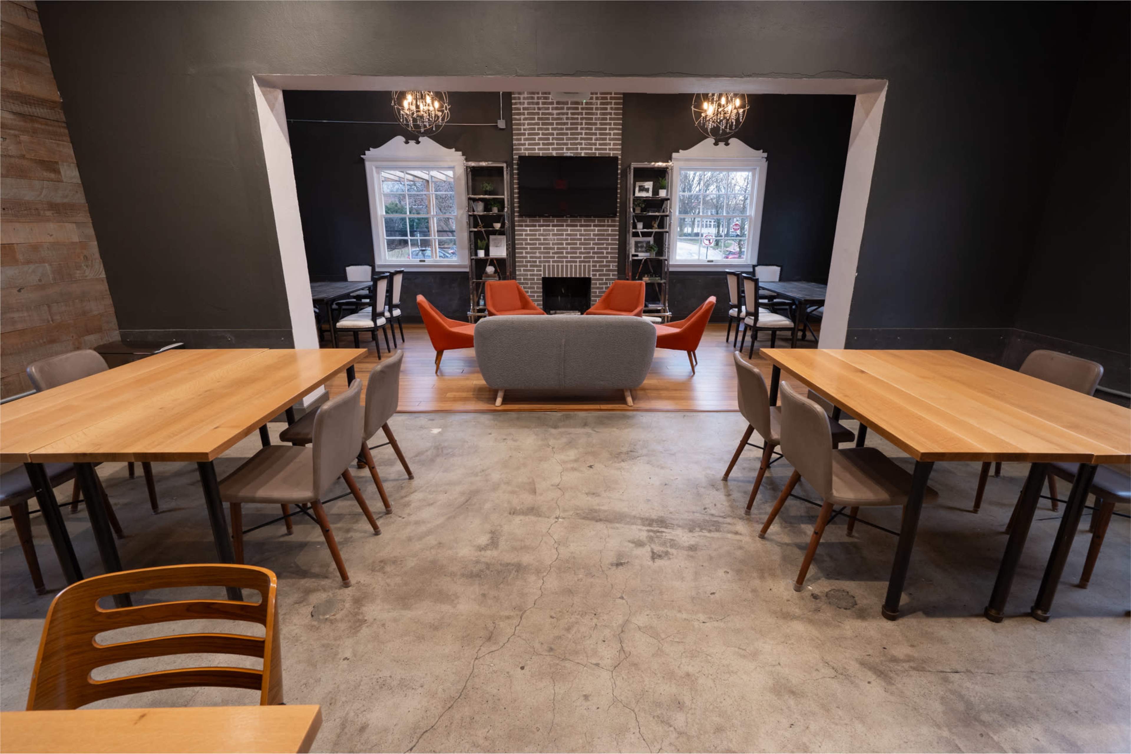 The image shows a modern interior of a cafe or meeting space with multiple wooden tables, gray and orange seating, and large windows allowing natural light.
