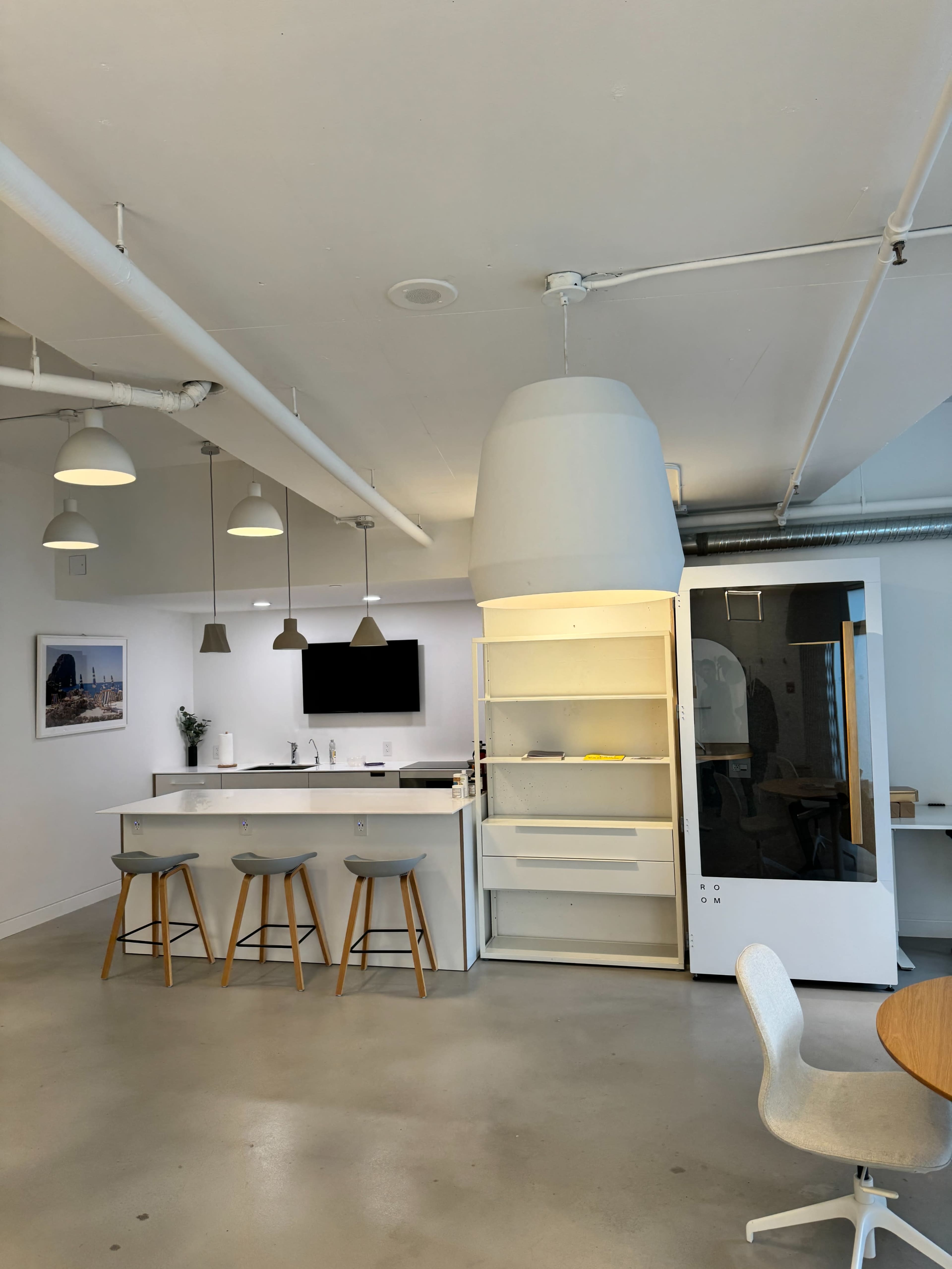 A modern office kitchen area with a counter, four stools, and a large white cabinet next to a seating area.