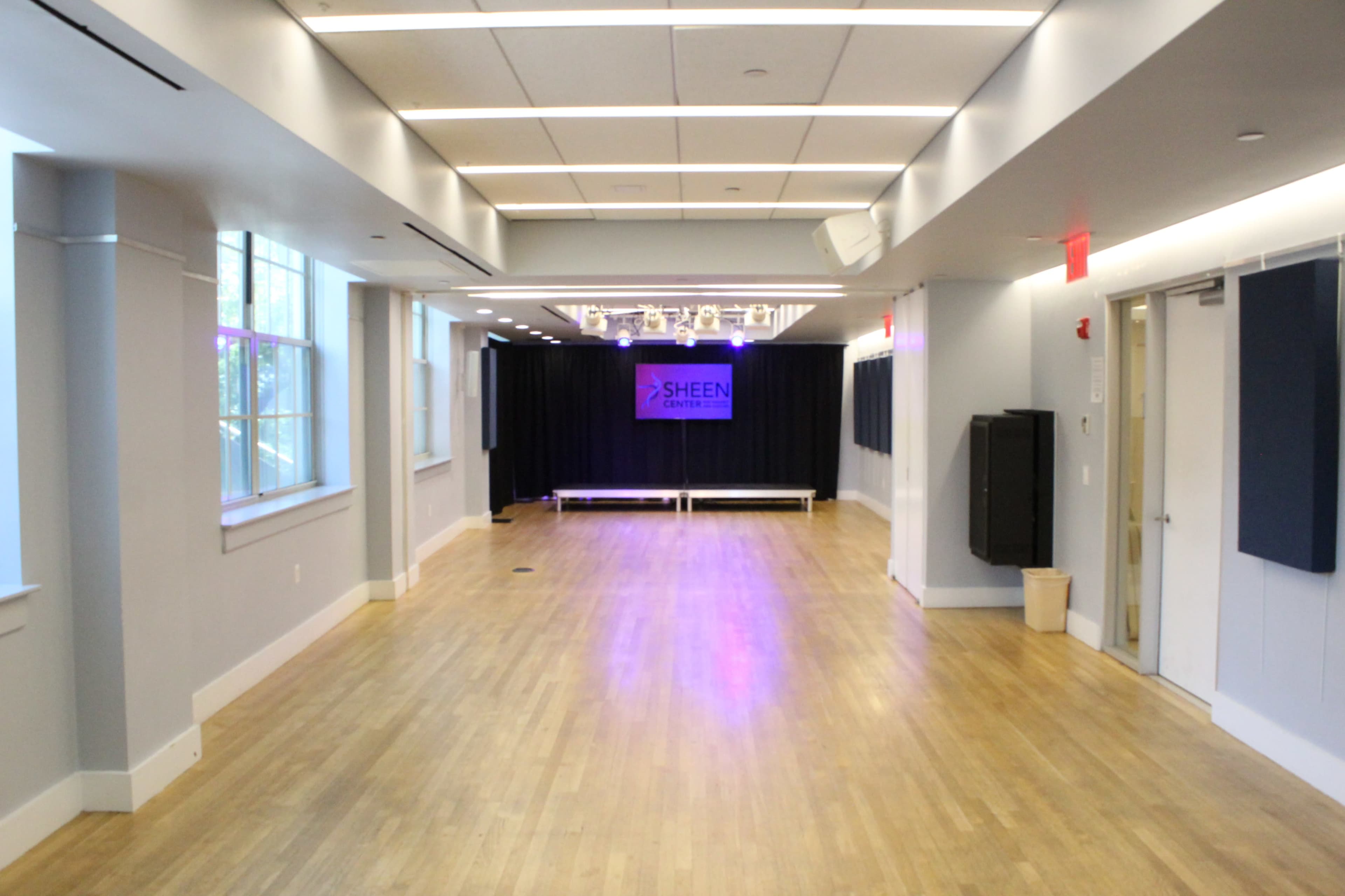 The image shows a spacious room with hardwood flooring, large windows on one side, and a performance stage with a backdrop at the far end.