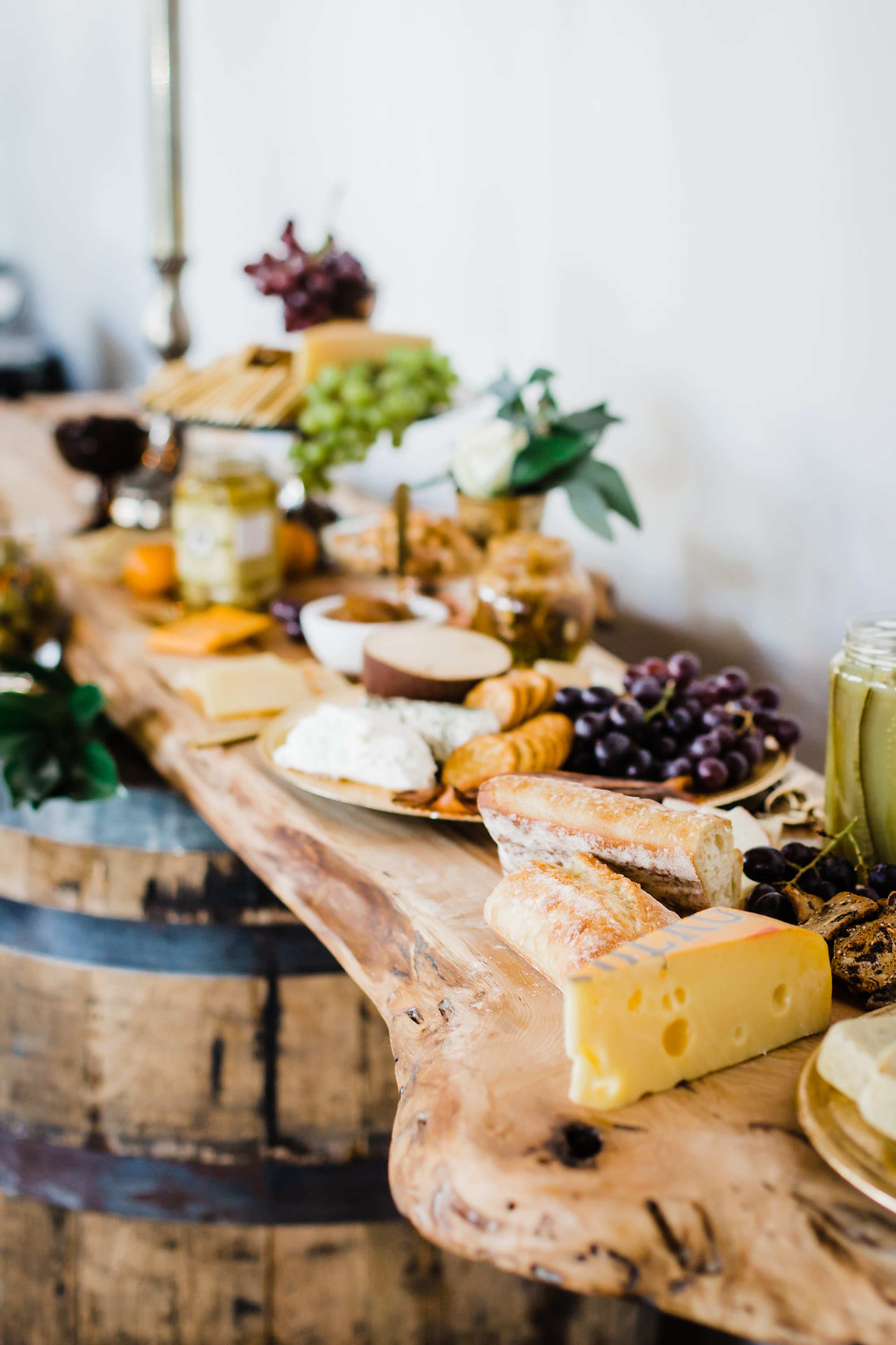 A long wooden table displays an assortment of cheeses, fruits, and spreads, arranged neatly for a gathering.