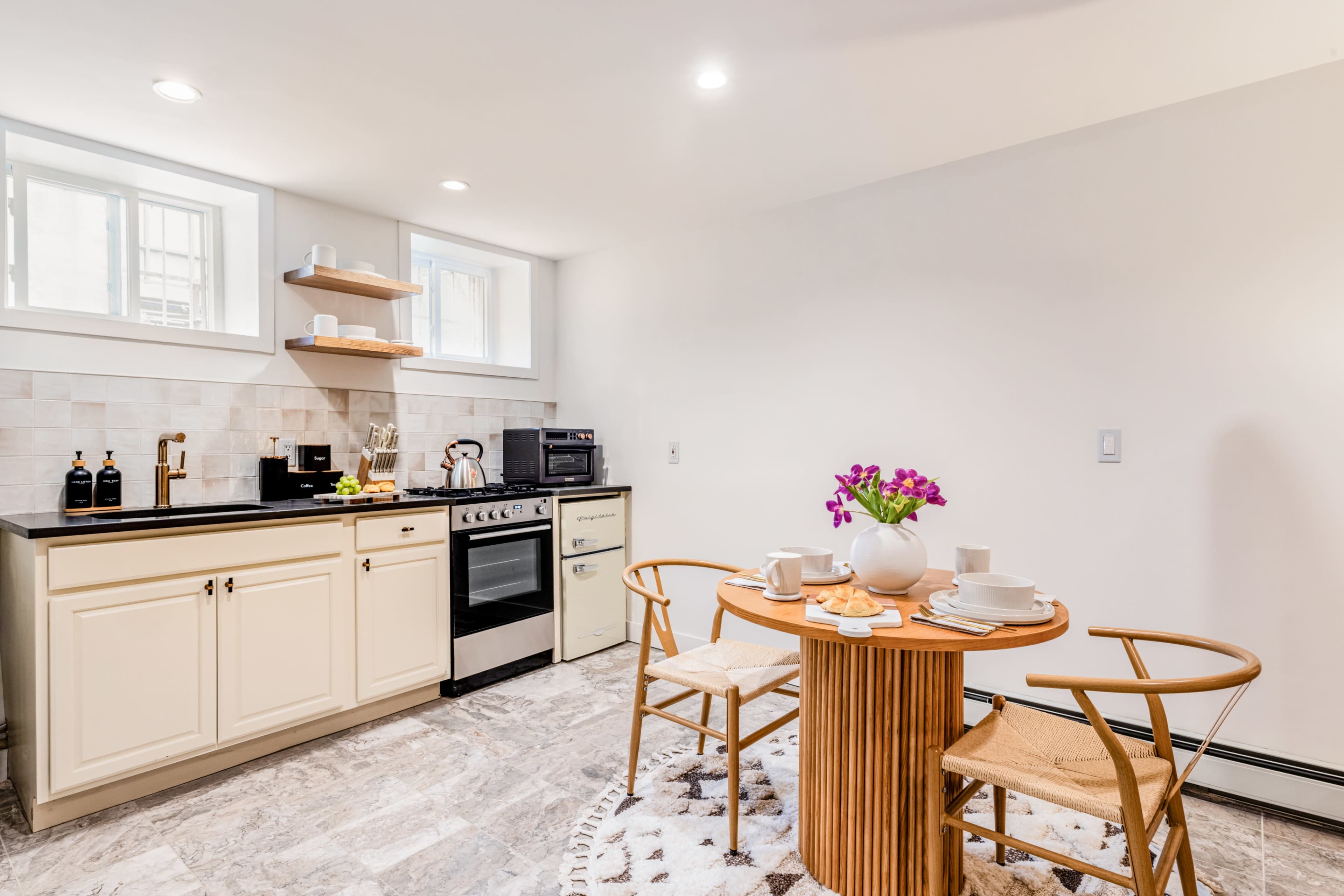 The image shows a small kitchen area featuring a table with two chairs, a stove, a microwave, and cabinets against a light-colored wall.