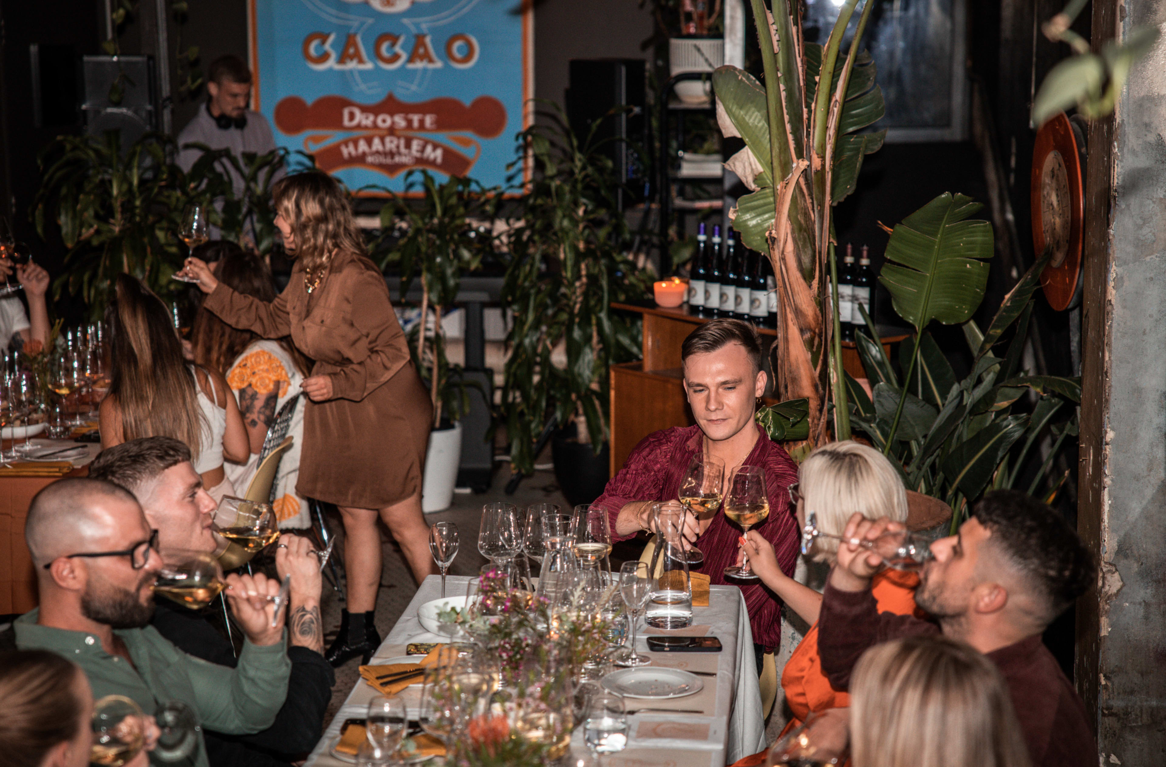 A lively group of people enjoys a dinner party surrounded by plants, with one woman serving drinks while others toast with glasses.