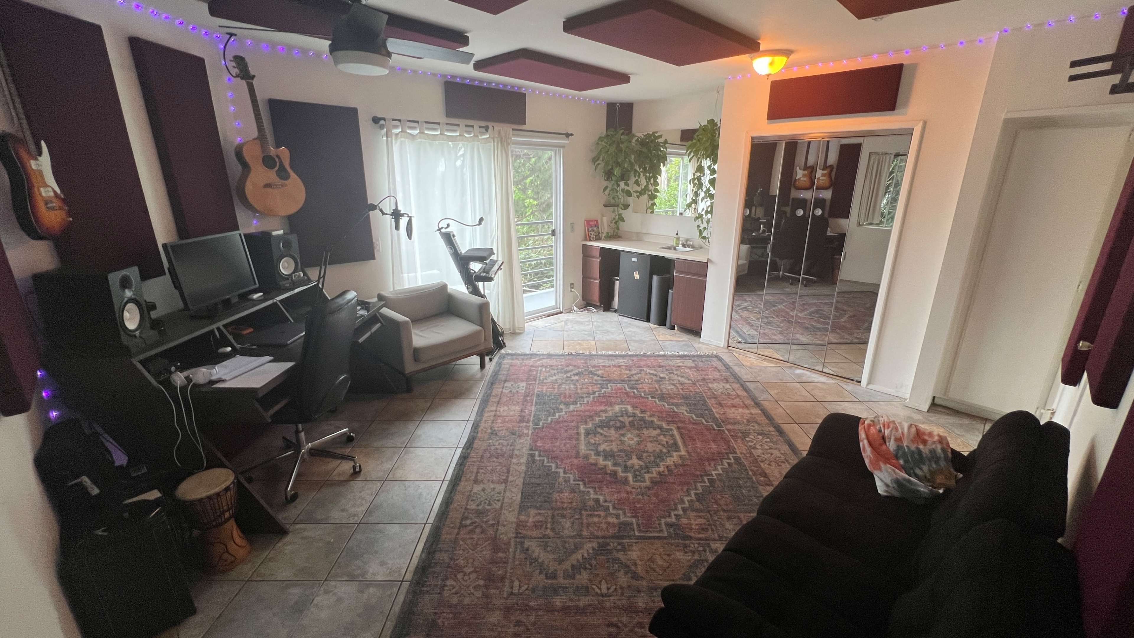 The image shows a music studio with a desk containing two computer monitors, an armchair, a black couch, and several guitars displayed on the walls.