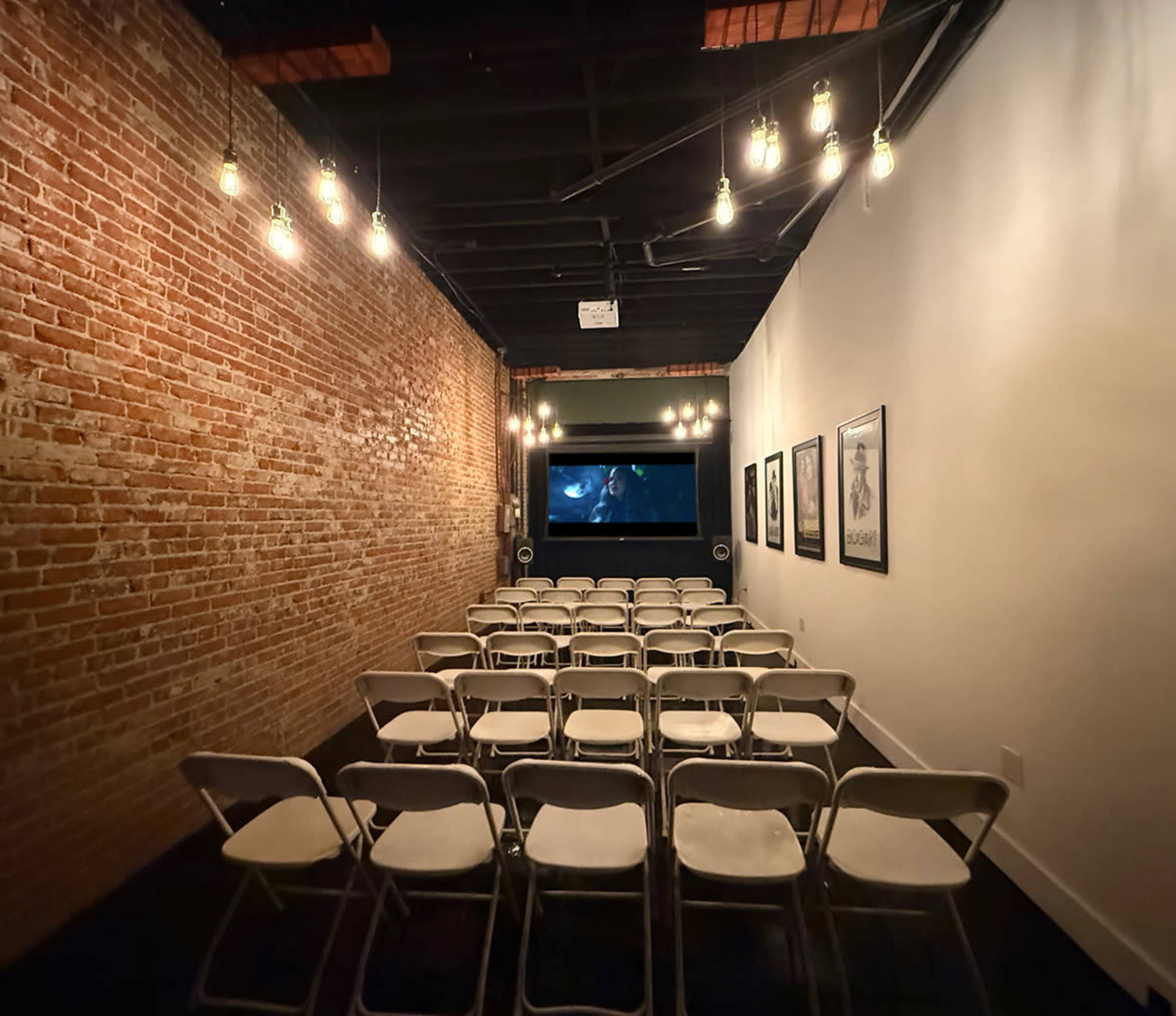 The image shows a narrow room with exposed brick walls, featuring rows of white folding chairs facing a large screen at the front, illuminated by hanging light bulbs.