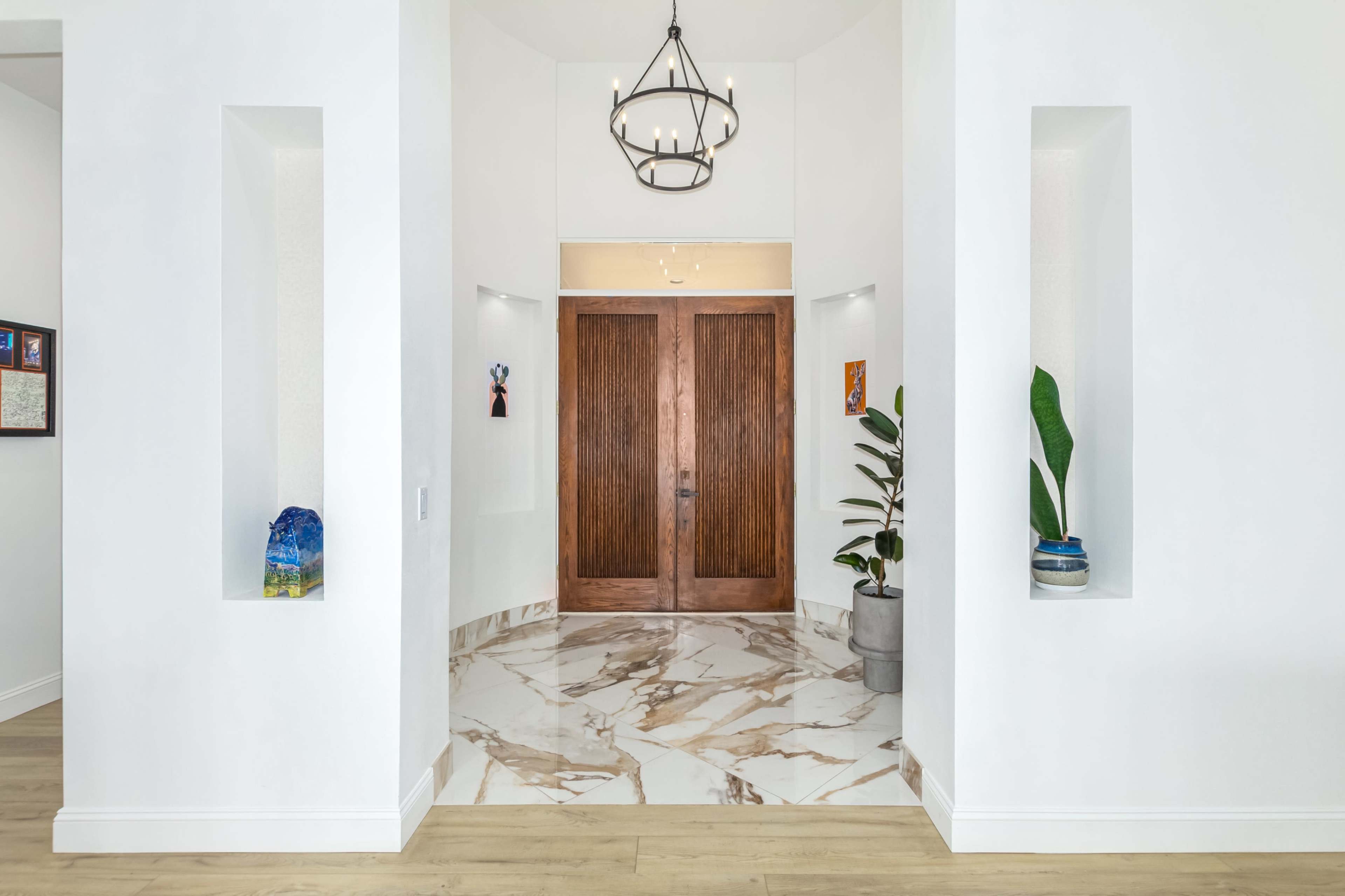 A spacious entryway with white walls, marble flooring, and large wooden doors, flanked by decorative niches and a chandelier above.
