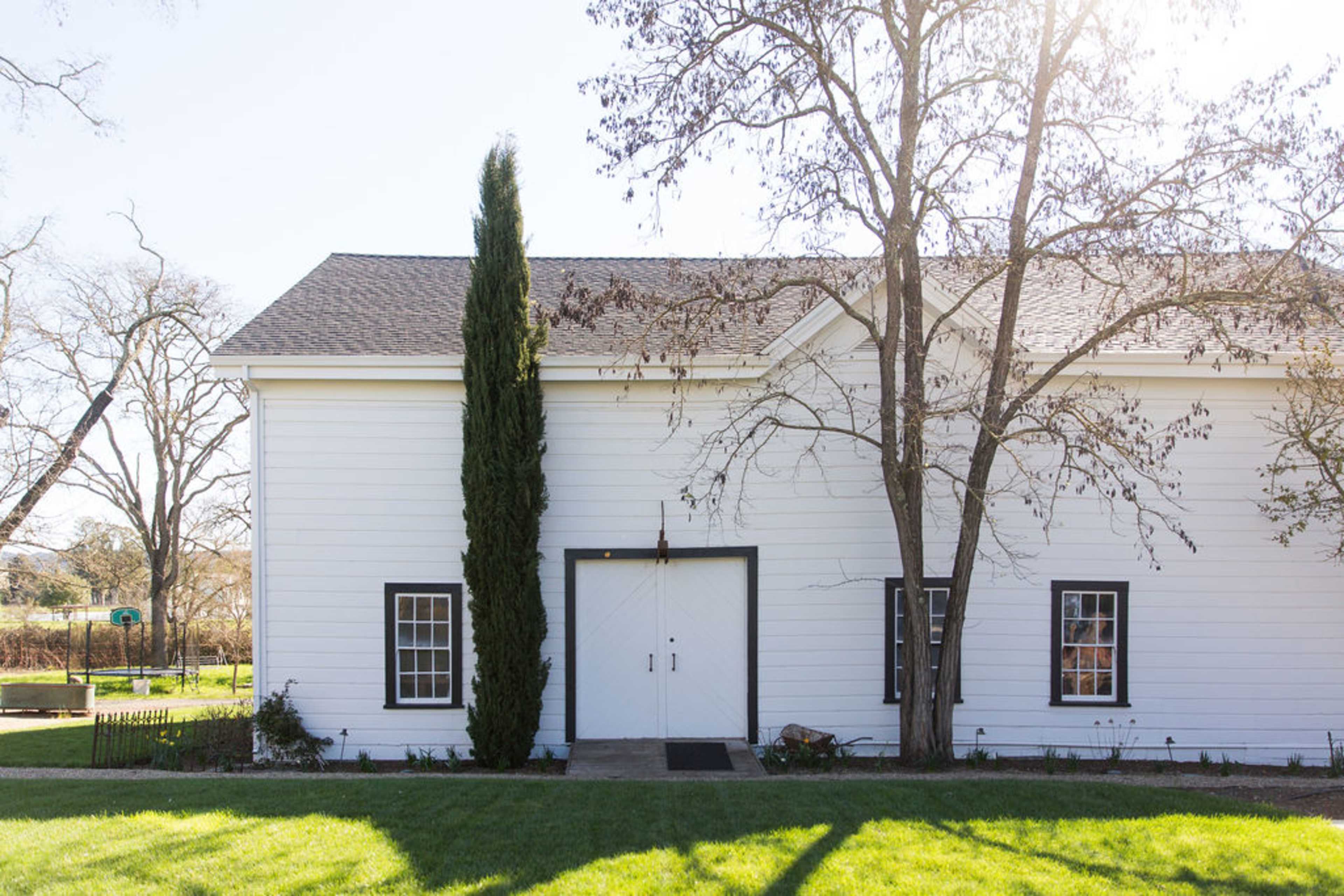 A white barn with a pair of double doors and two windows is set against a backdrop of trees and green grass.
