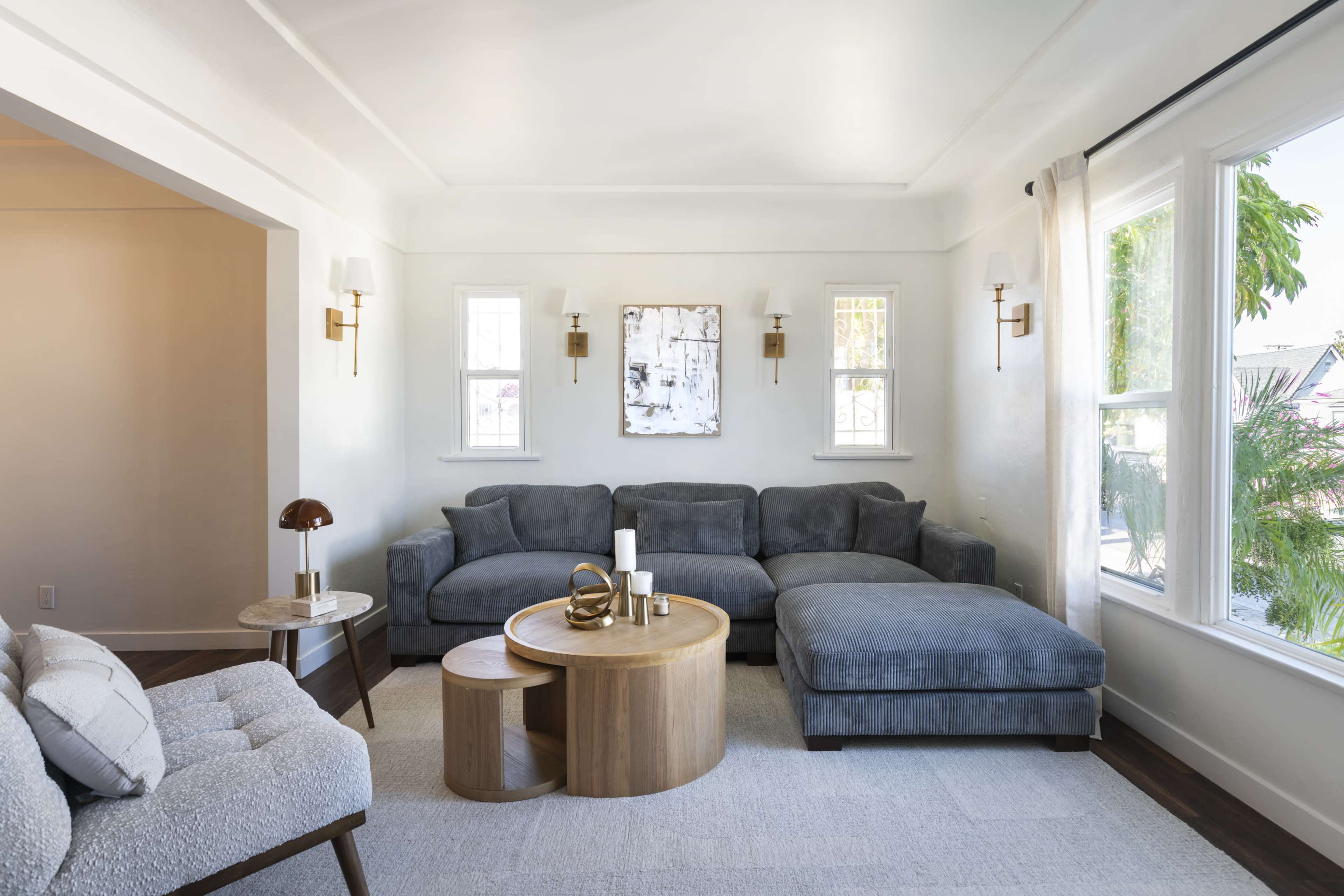 The image shows a modern living room featuring a gray sectional sofa, a round wooden coffee table, and large windows that allow natural light to fill the space.
