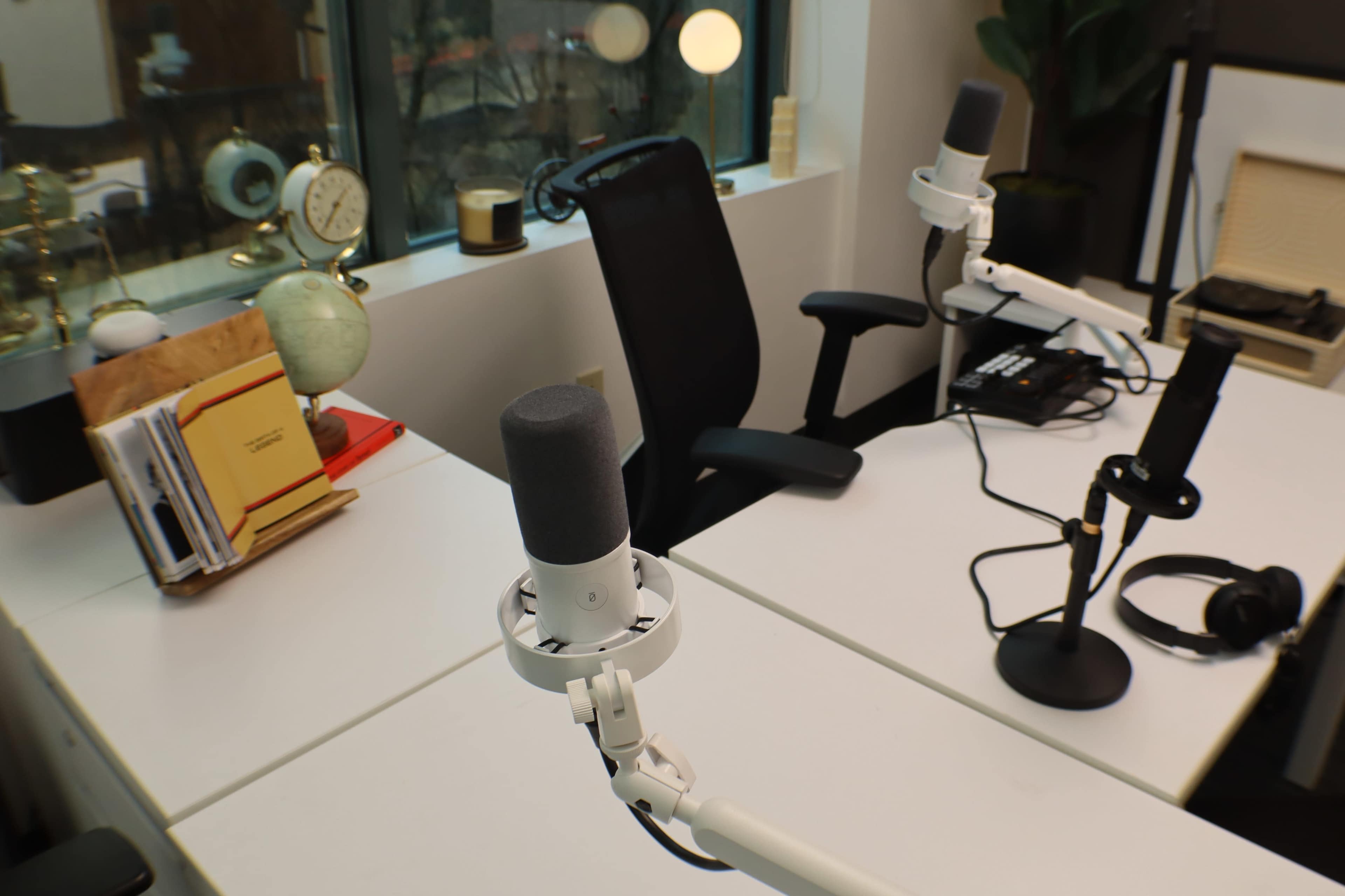 The image shows a podcast recording workspace with two microphones mounted on adjustable arms, a chair, and various decorative items on a desk near a window.