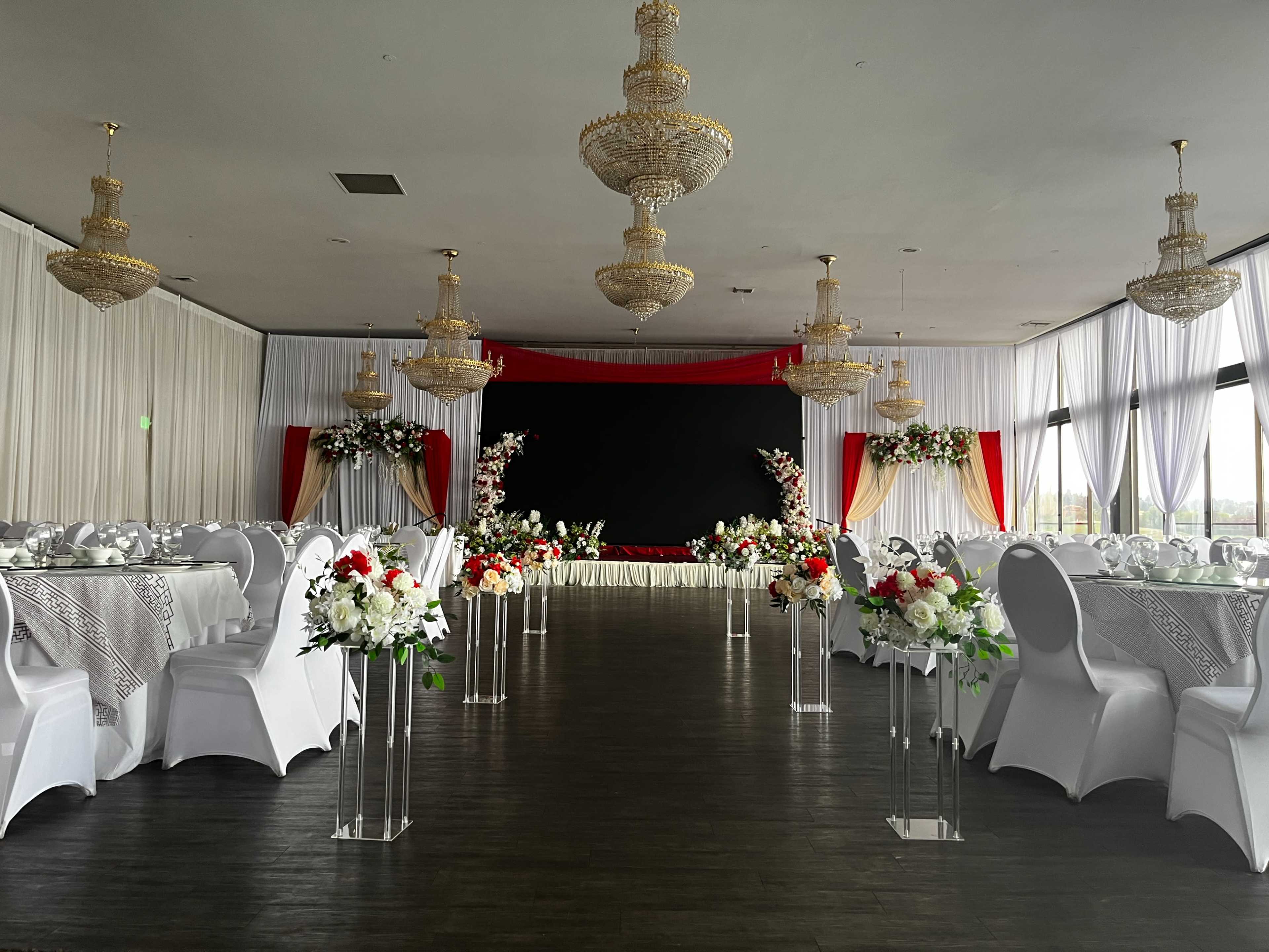 The image shows a banquet hall beautifully arranged with white tables adorned with floral centerpieces and elegant chandeliers hanging from the ceiling.