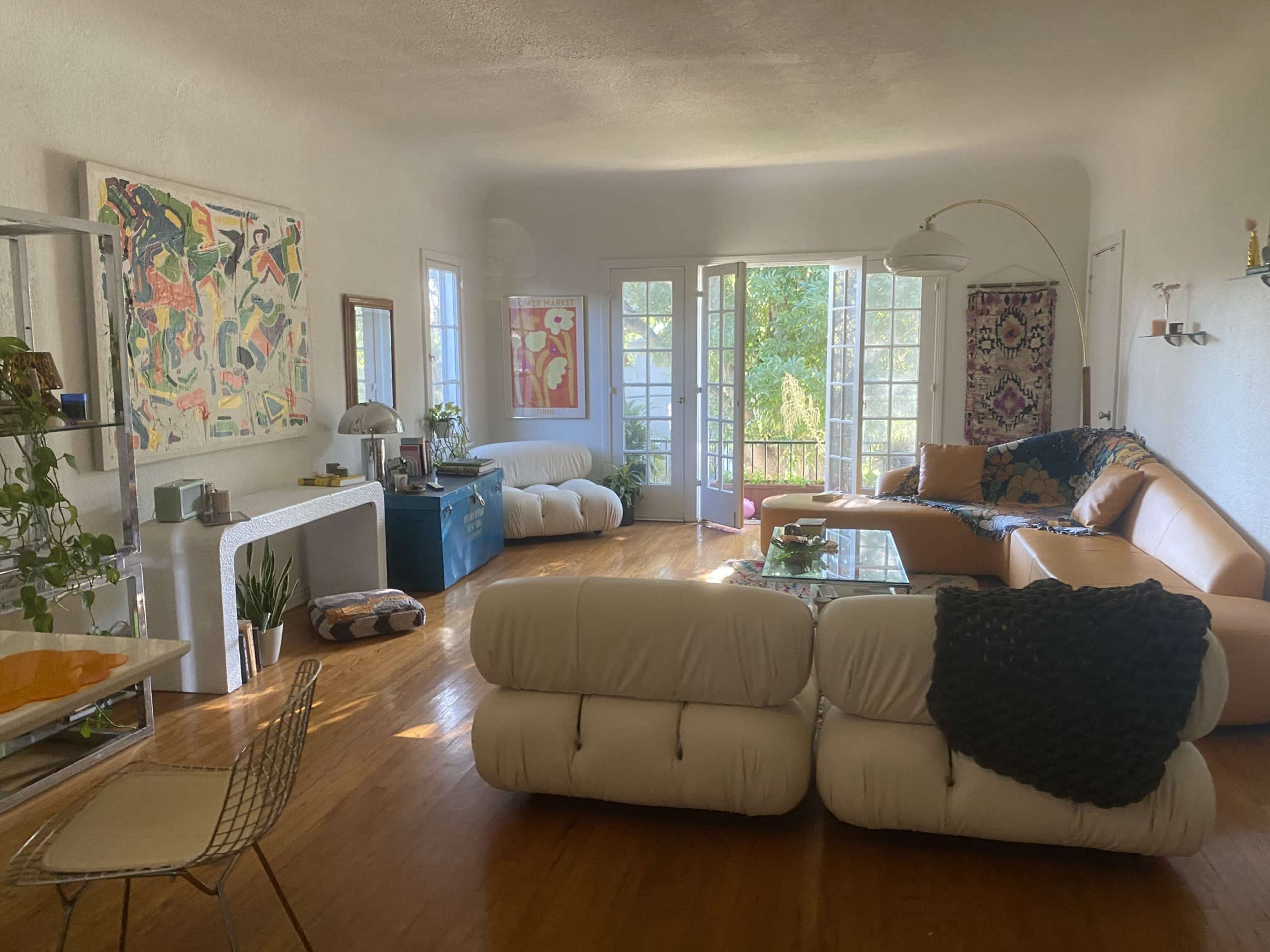 A modern living room with light-colored walls, multiple seating areas, large windows opening to a patio, and various pieces of art displayed on the walls.