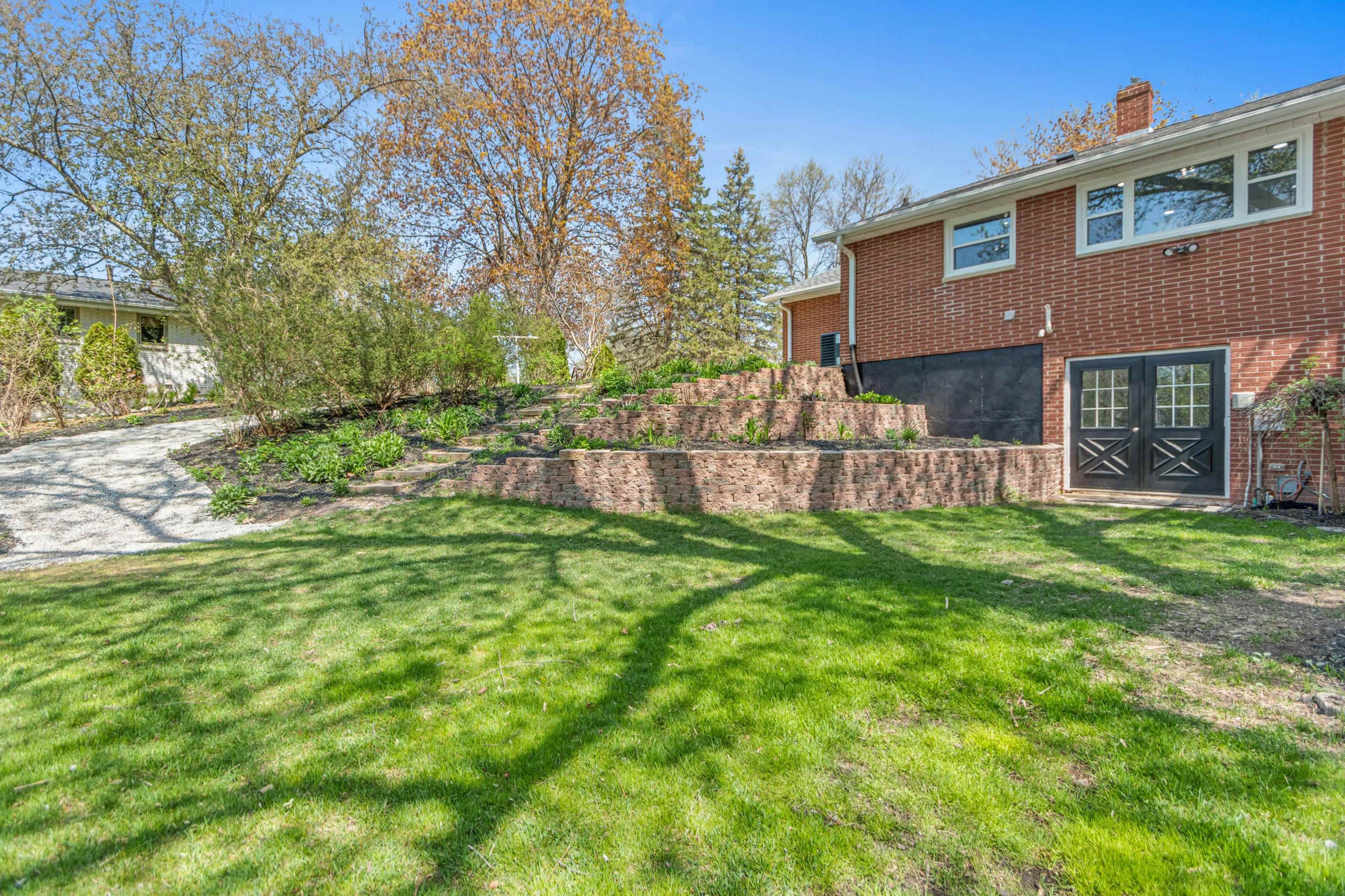 A landscaped yard with tiered brick retaining walls leading up to a brick house, surrounded by trees and a grassy area.