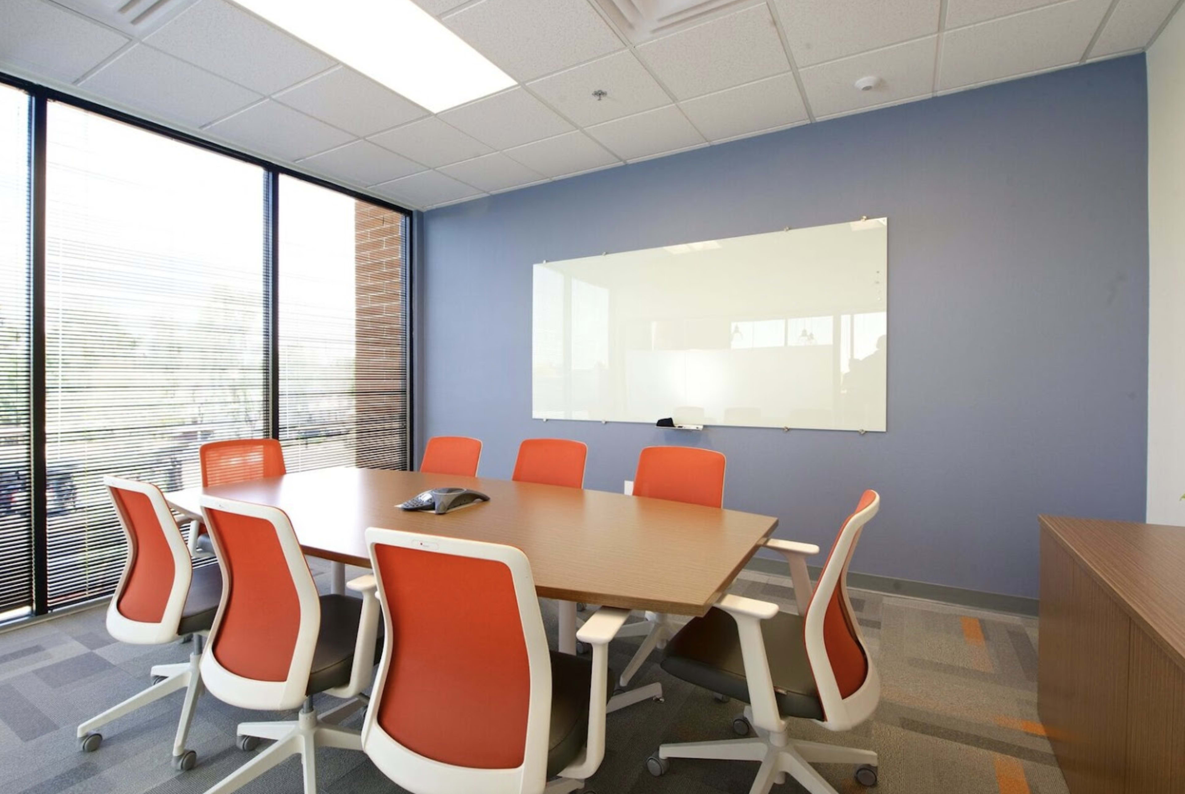 A modern conference room featuring a large wooden table surrounded by eight orange-accented chairs, with a glass whiteboard on the wall and windows allowing natural light.