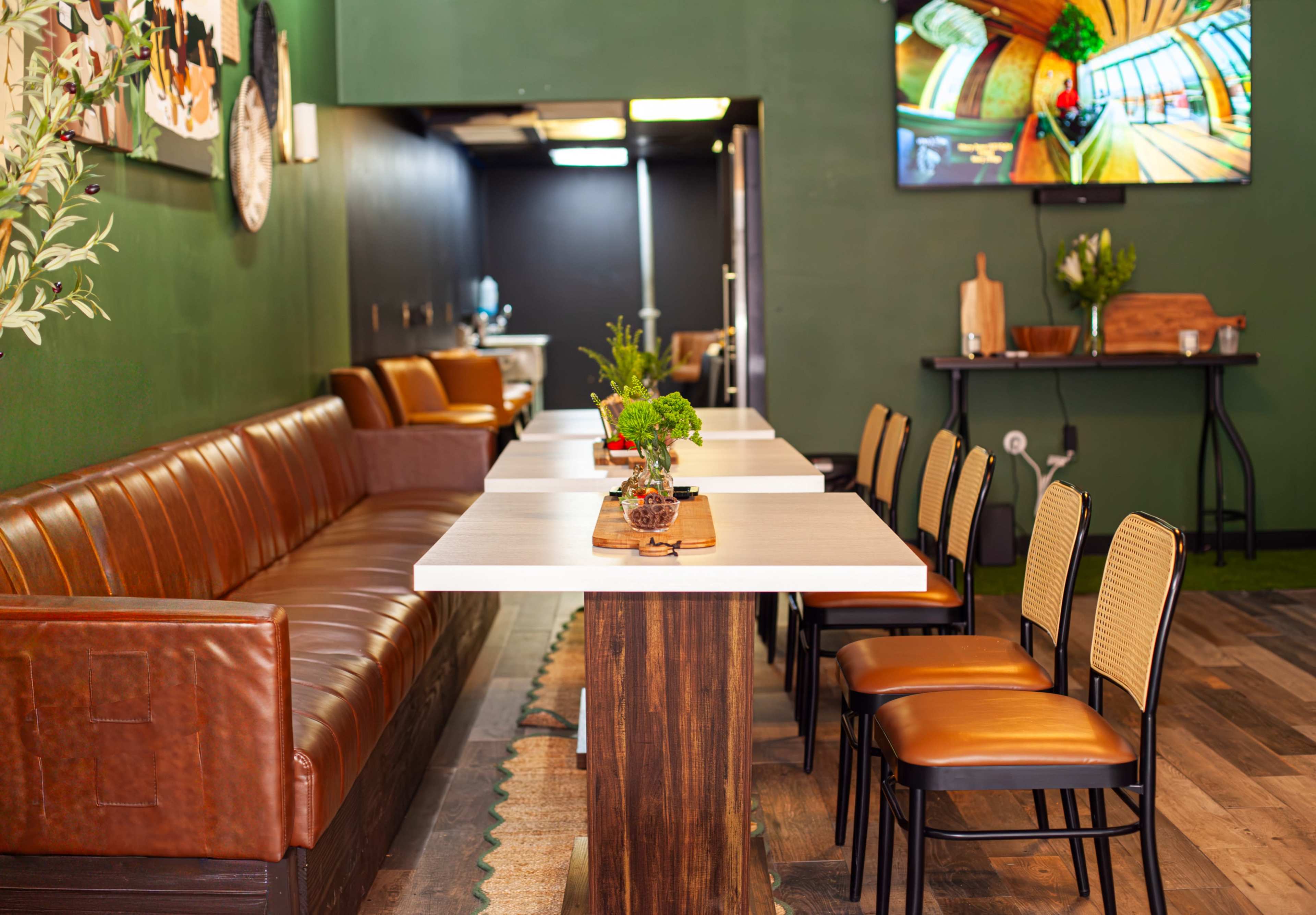 The image shows a cozy indoor space with a brown leather sofa, a long table with potted plants, and several wooden chairs arranged around the table, all set against green walls and equipped with a wall-mounted TV.